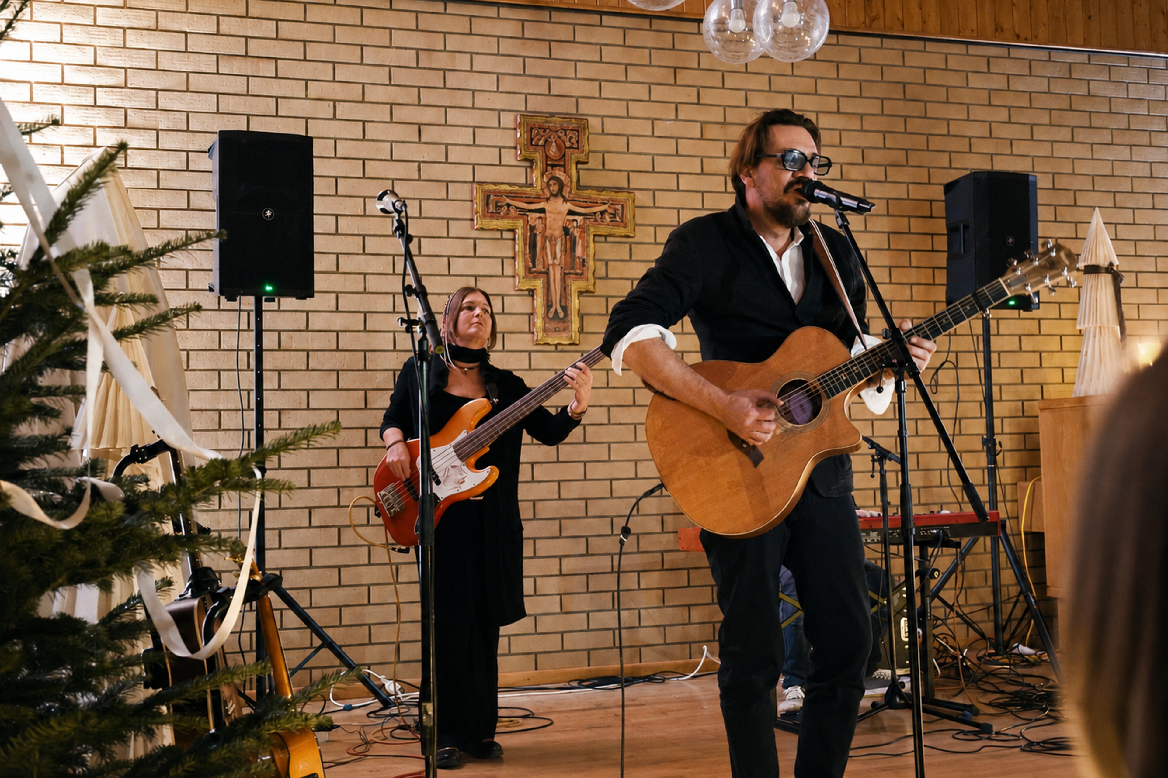 Man wearing glasses sings into microphone while playing acoustic guitar, woman plays bass guitar behind him, both in front of brick wall with cross and speakers in wood-paneled room
