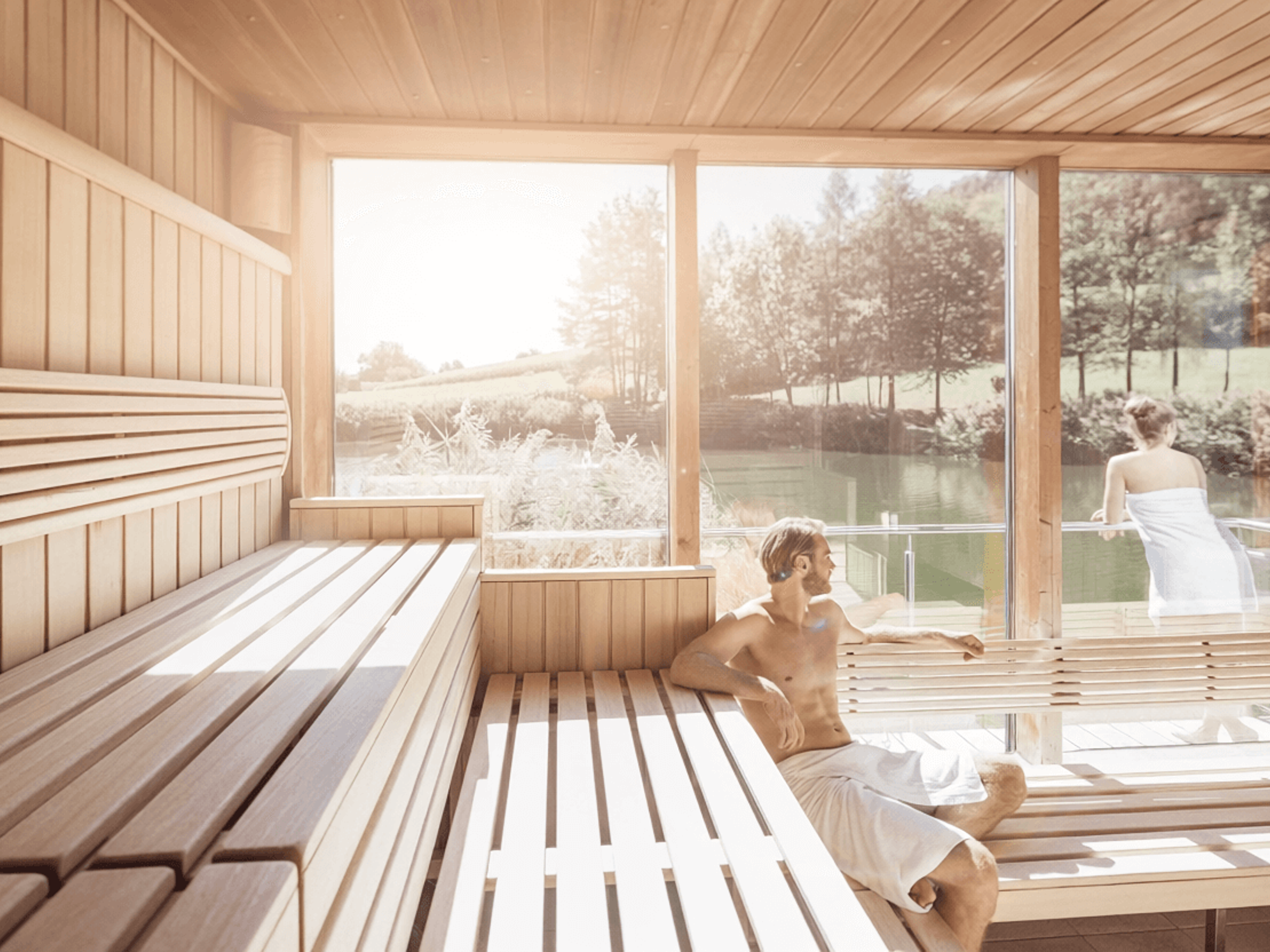 Innenraum einer Sauna mit Holzbänken und großen Fenstern, durch die eine grüne Landschaft sichtbar ist, zwei Personen mit Handtüchern sitzen und stehen auf den Bänken