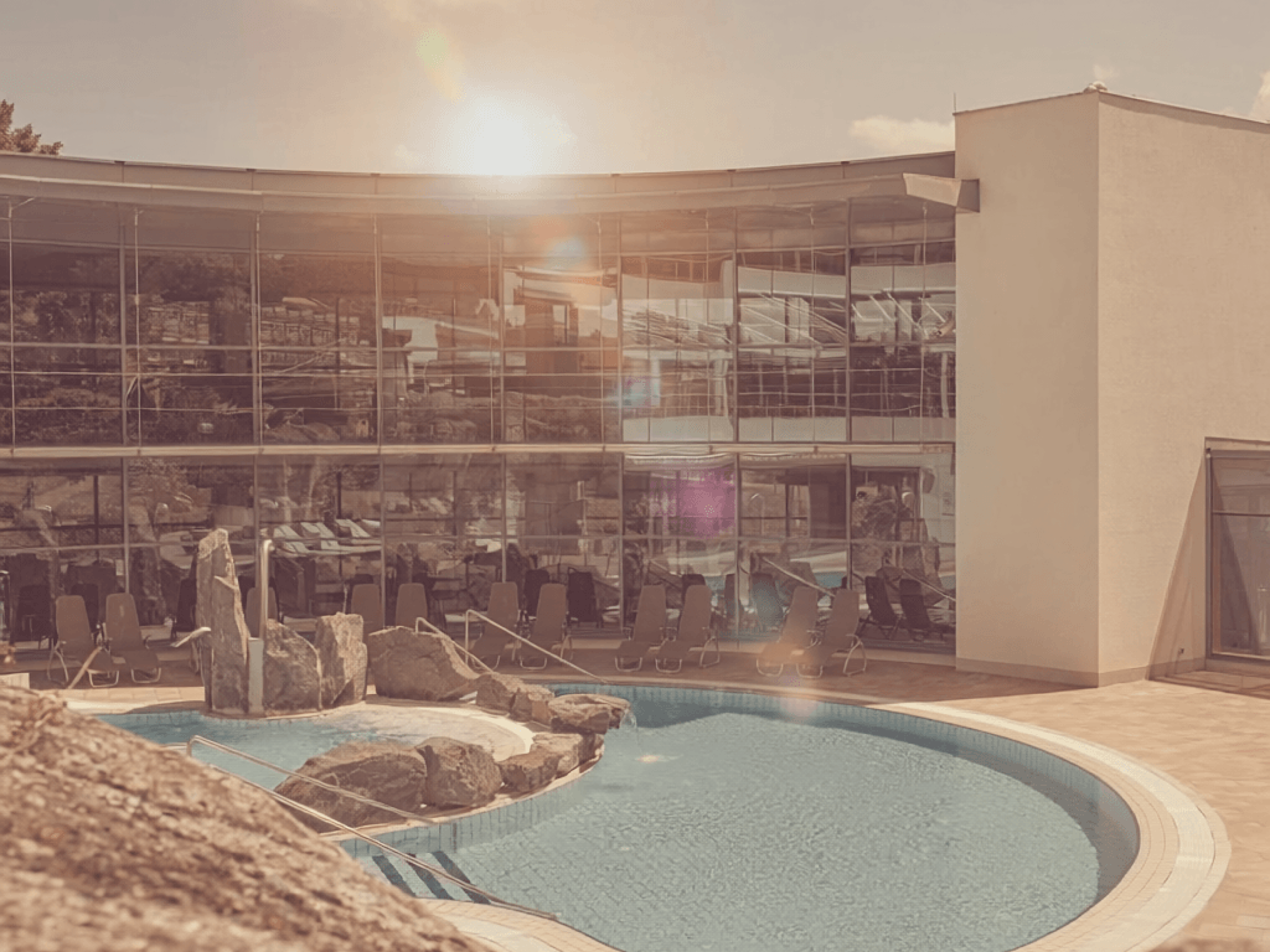 Outdoor pool with rock island in front of a modern building with large glass facade under blue sky