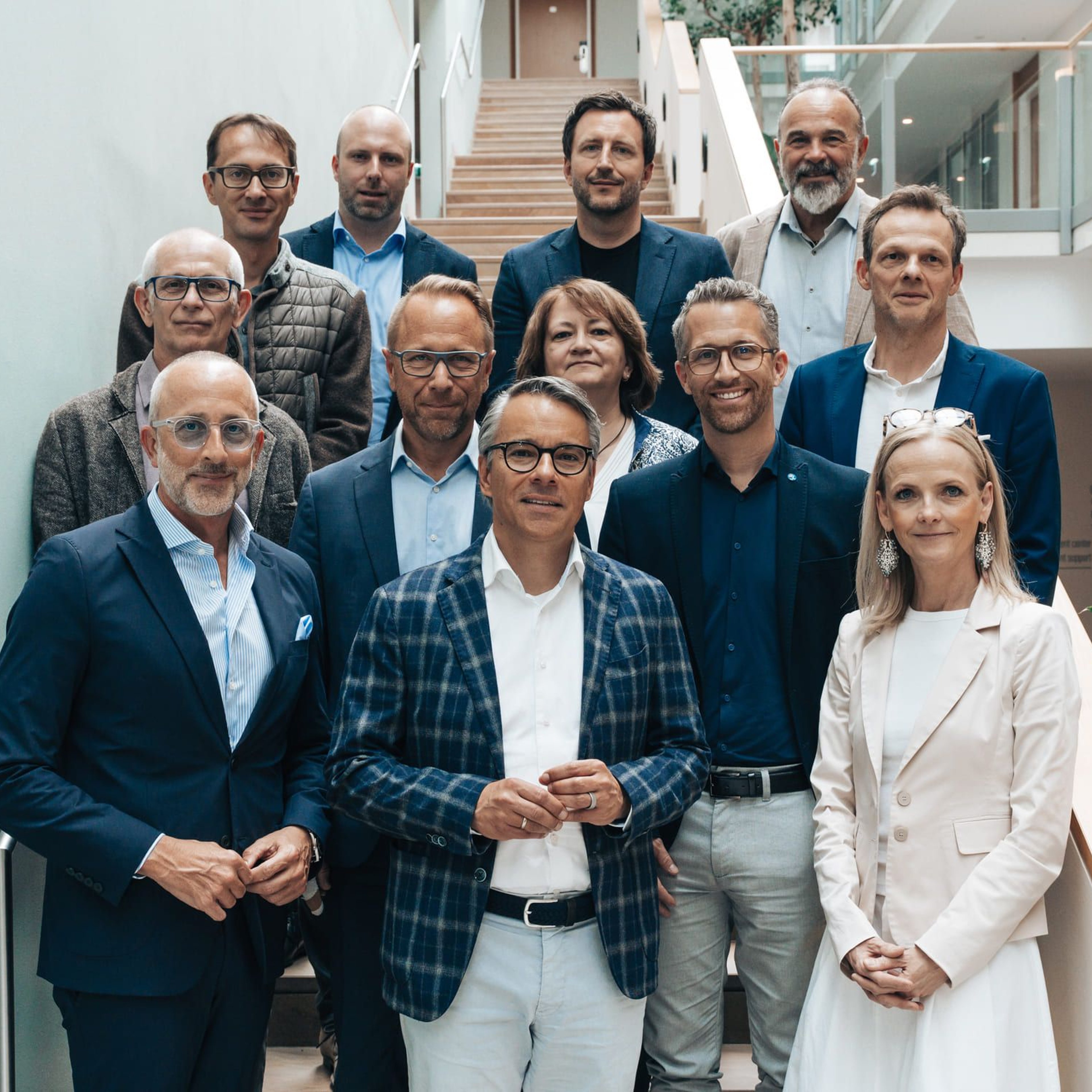 Zwölf Personen in formeller Kleidung stehen in einem modernen Innenraum mit Treppe und Glasgeländer, darunter neun Männer und zwei Frauen.
