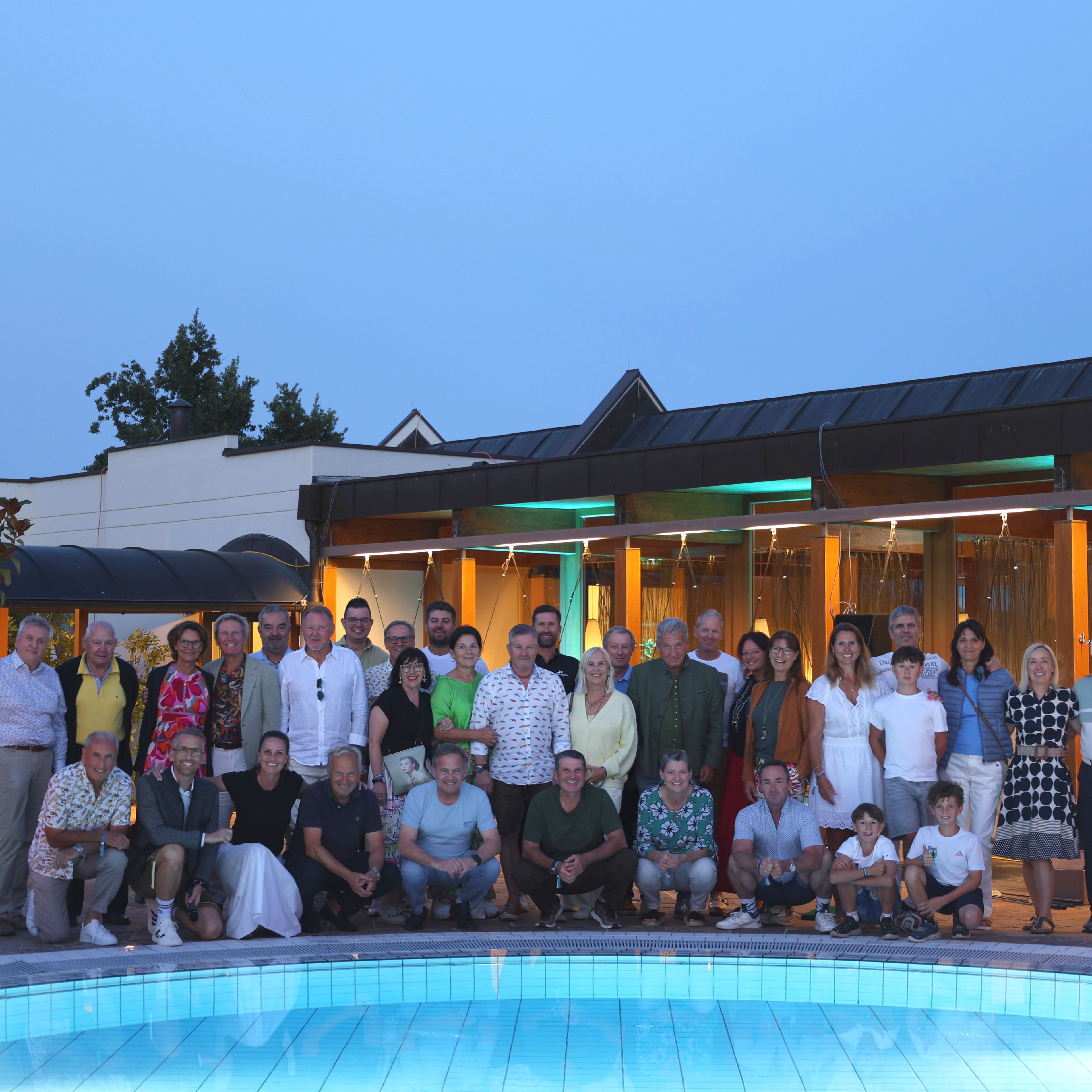 Große Gruppe von Menschen unterschiedlichen Alters steht und sitzt vor einem beleuchteten Gebäude neben einem Pool bei Abenddämmerung