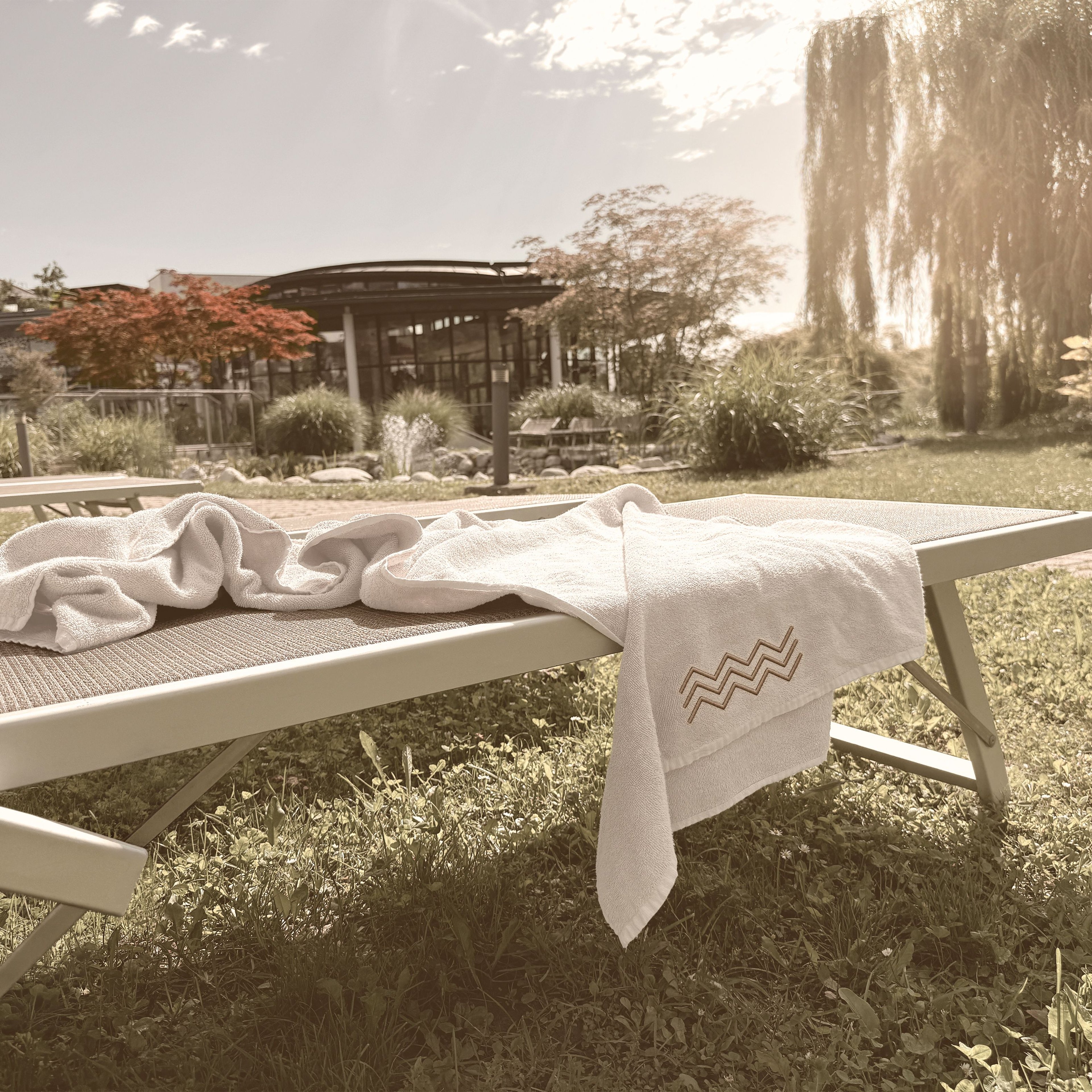 Lounge chair with two white towels on grass in front of a modern building and trees