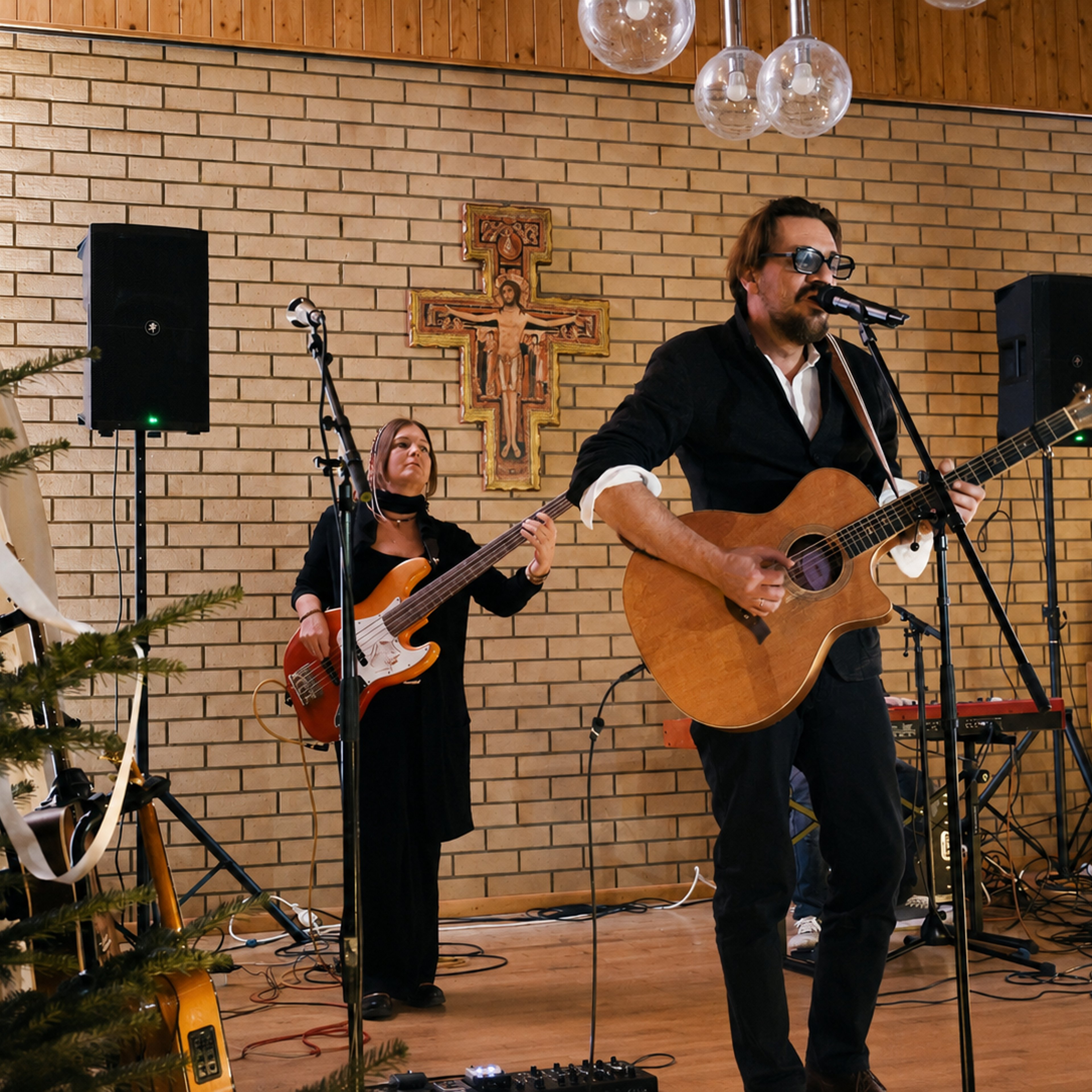 Mann mit Gitarre und Brille singt vor Mikrofon, Frau spielt Bassgitarre, beide vor einer Wand mit Kreuz und Lautsprechern in einem Raum mit Holzverkleidung