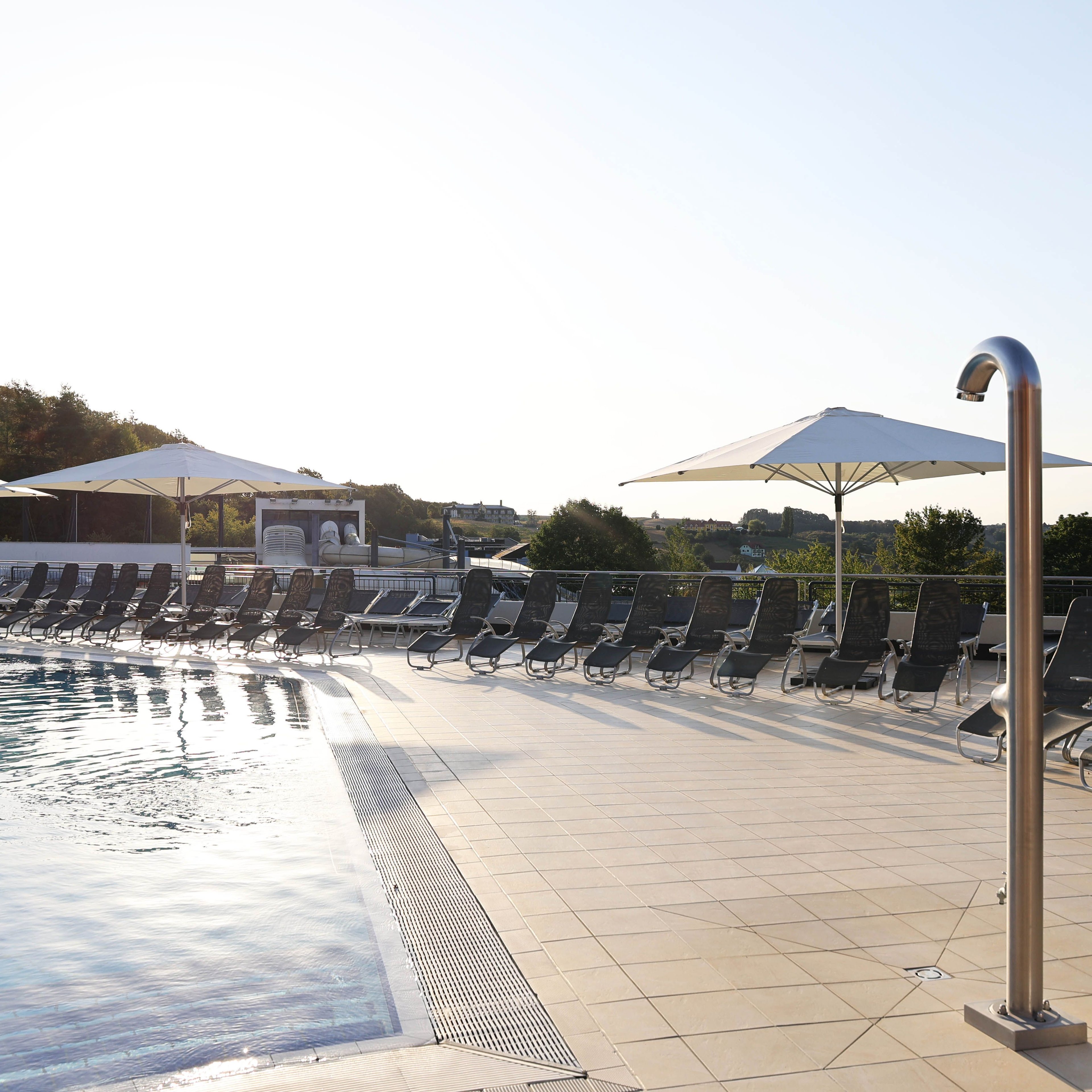 Außenterrasse eines Thermenbads mit Liegestühlen, Sonnenschirmen, einem Pool und einer Außendusche auf hellen Fliesen