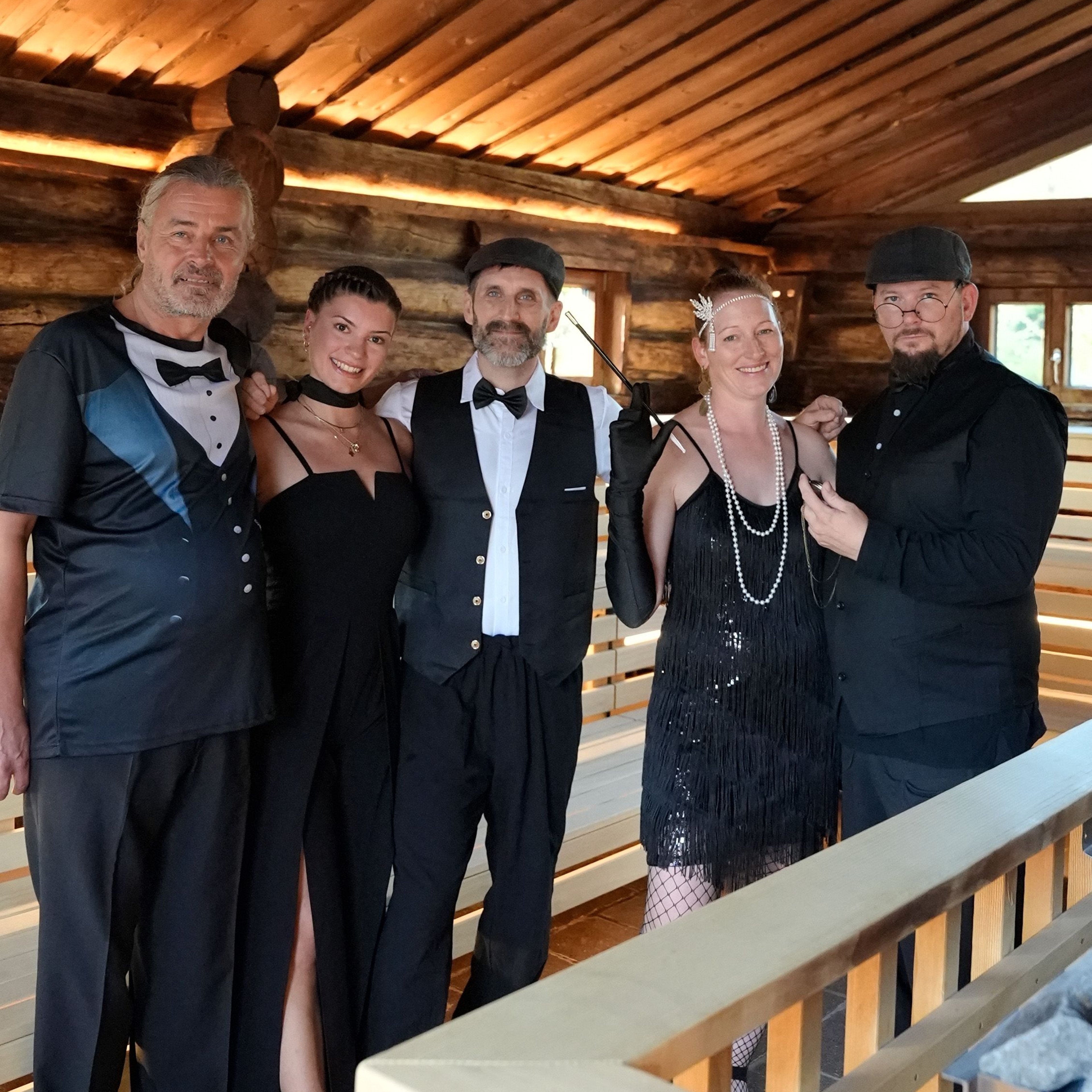 Fünf Personen in 1920er-Jahre-Kleidung stehen in einem Raum mit Holzwänden und -decke, zwei Frauen tragen elegante Kleider, drei Männer Anzüge und Fliegen.