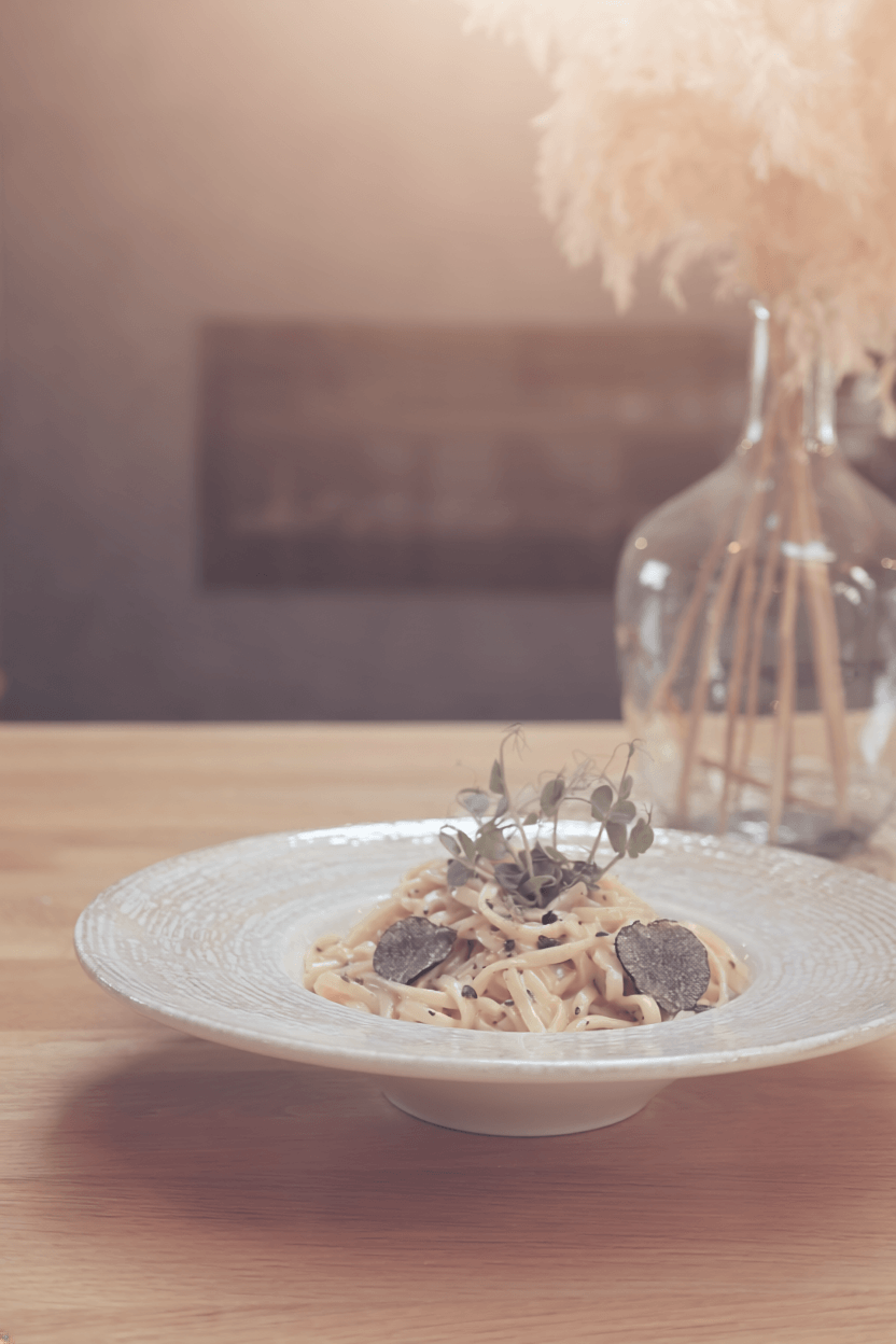 Plate of pasta garnished with truffle slices and fresh herbs on a wooden table in front of a vase of dried flowers.