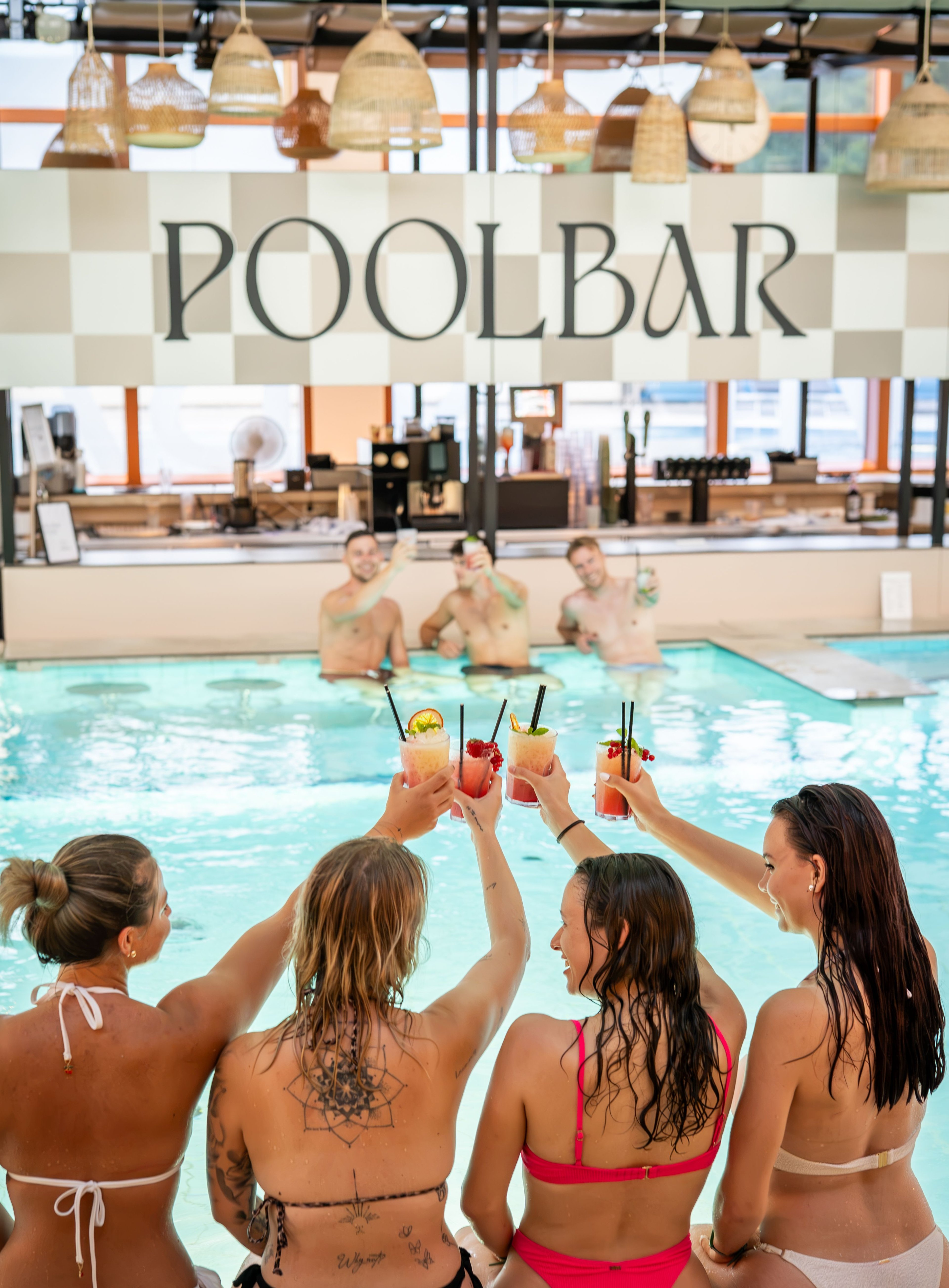 Vier Frauen in Badebekleidung sitzen am Beckenrand und stoßen mit bunten Cocktails auf drei Männer im Wasser an, im Hintergrund ein Schild mit der Aufschrift 'POOLBAR'