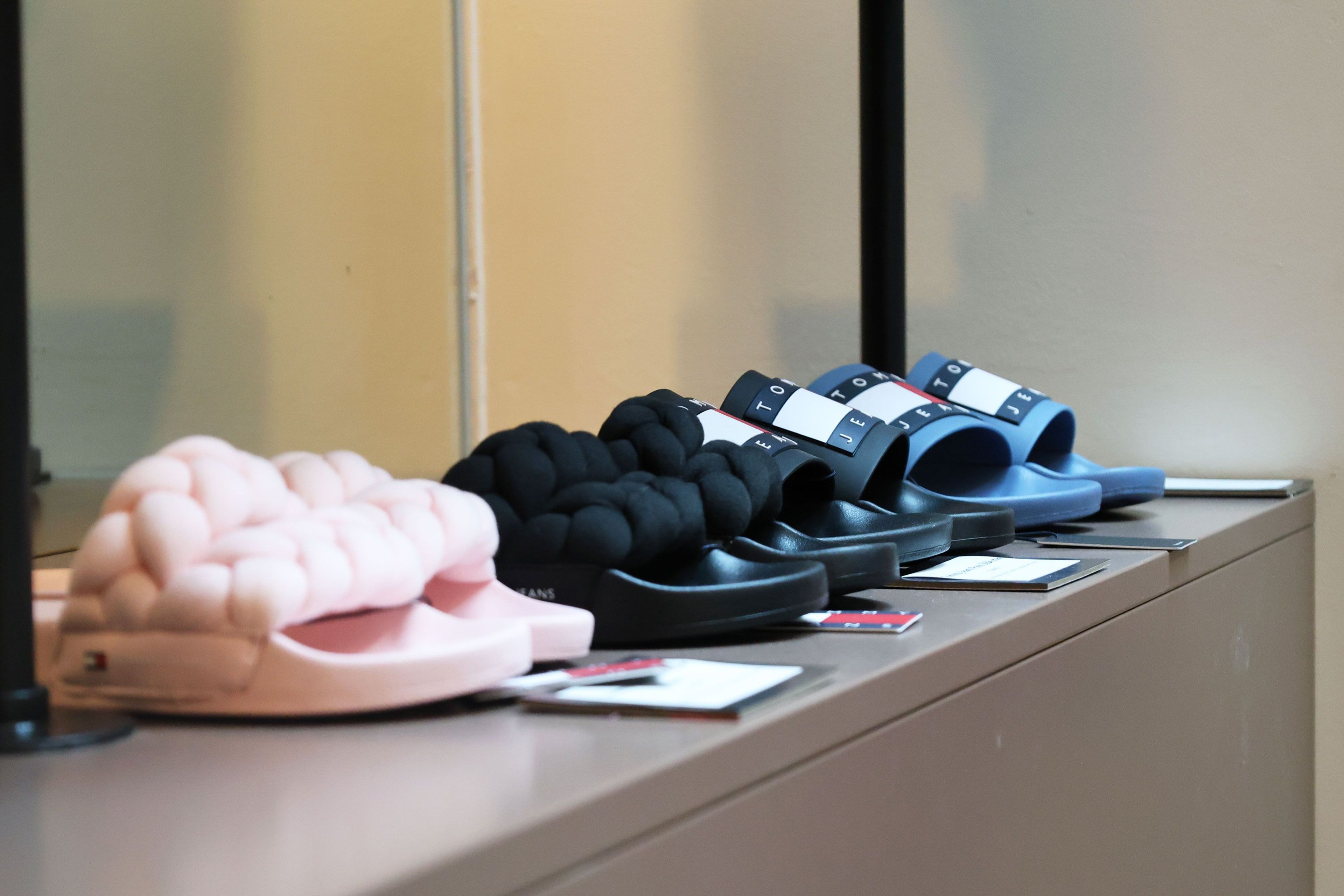 Four pairs of slippers in pastel pink, black, and blue with padded, knot-like straps on a shelf.
