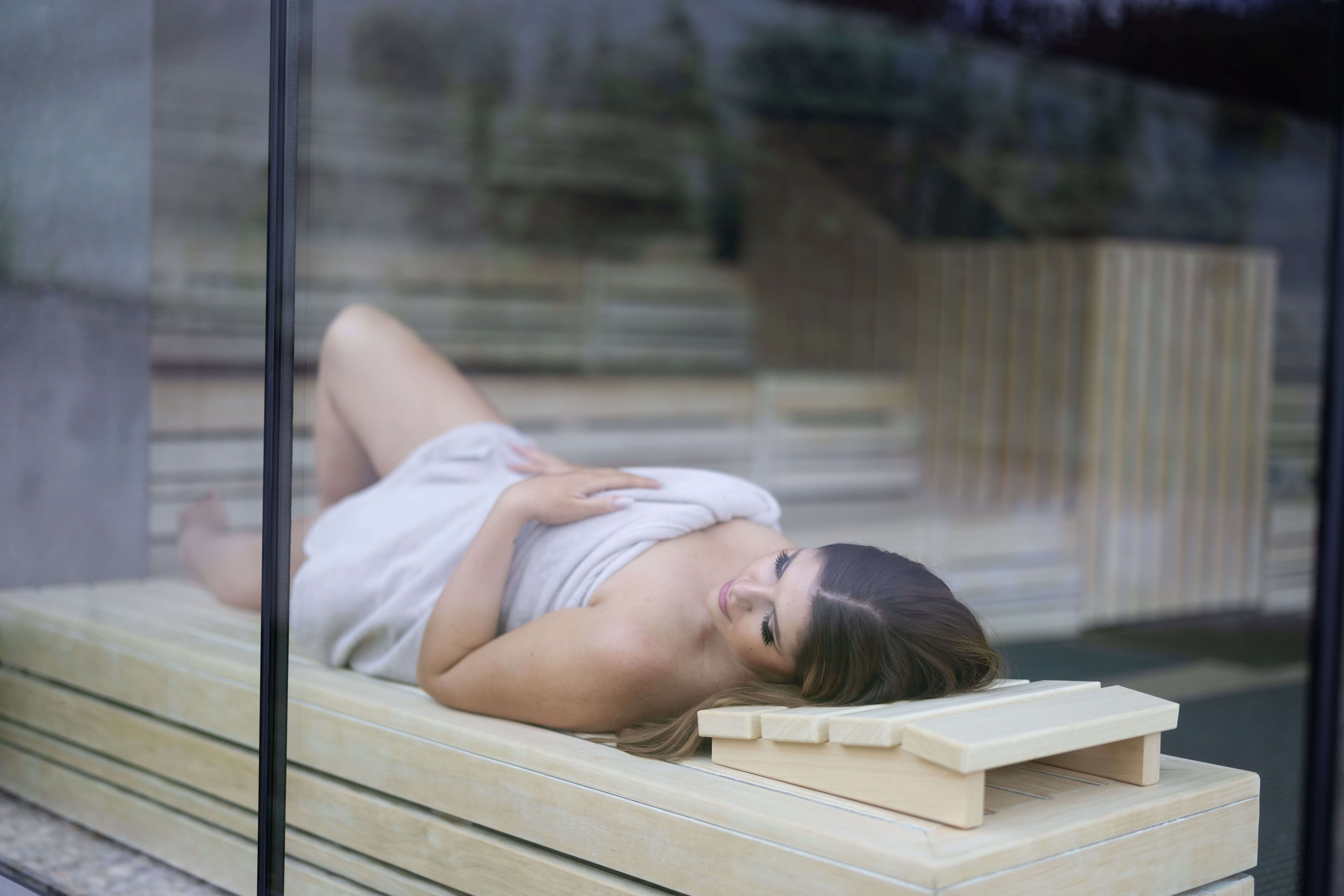 Woman lying on a brown wooden bench in the sauna, eyes closed and enjoying the warmth.
