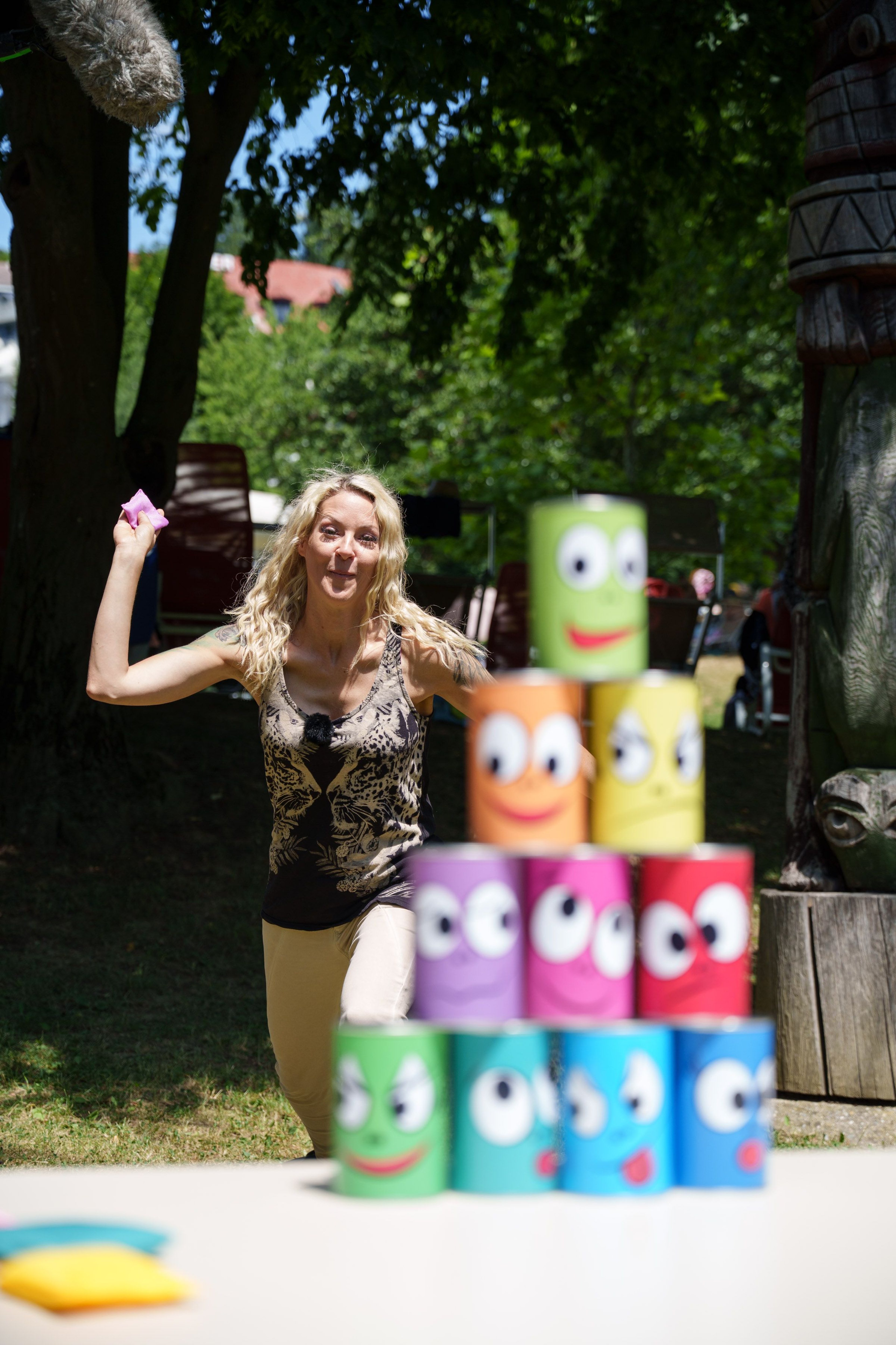 Woman throwing a small object with her right hand at a tower of colorful cans with faces outdoors under trees