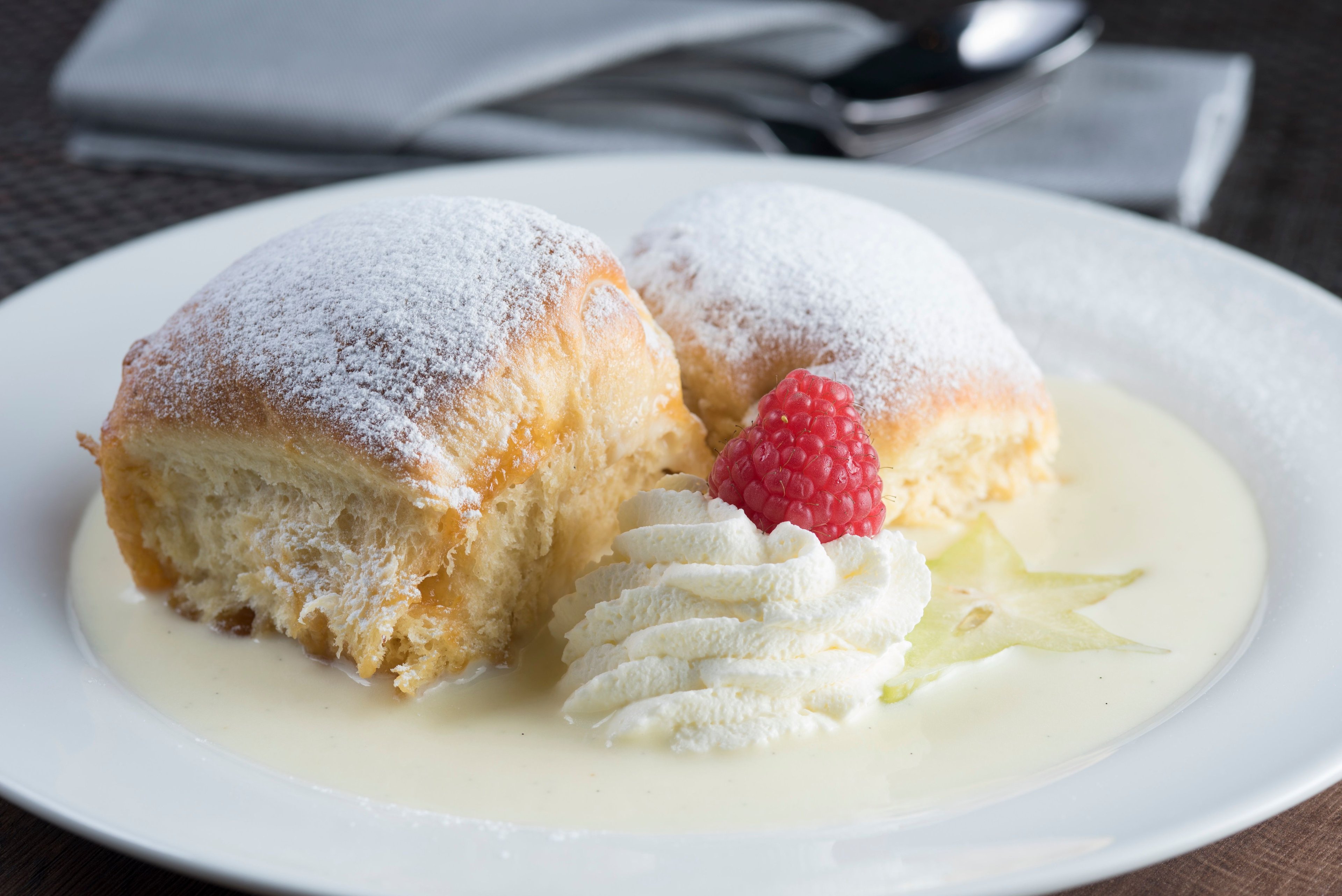 Two powdered sugar dusted Buchteln on a white plate with vanilla sauce, whipped cream, and a raspberry