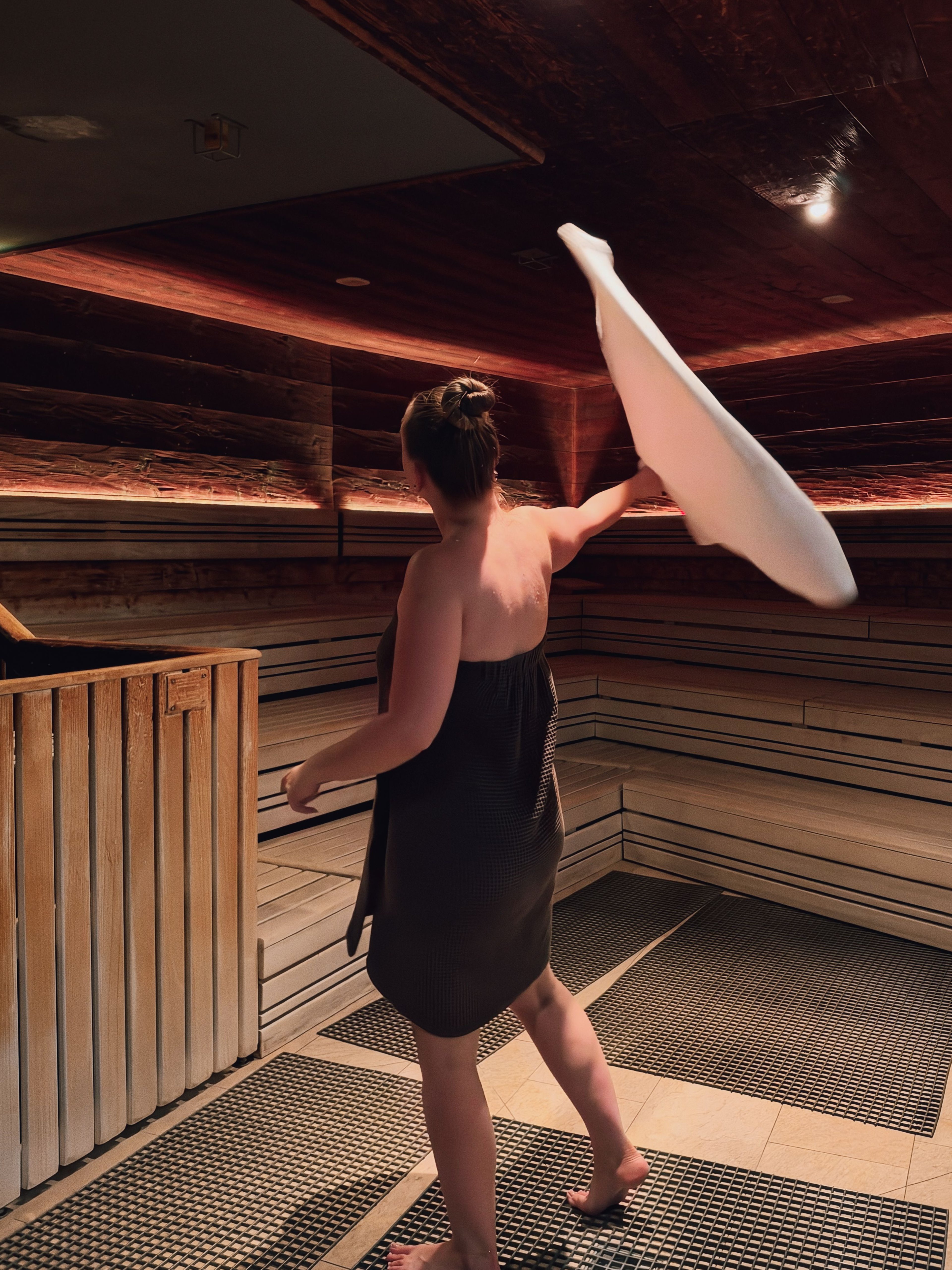 Frau mit hochgestecktem Haar und schwarzem Handtuch wirbelt ein weißes Handtuch in einer Sauna mit Holzverkleidung