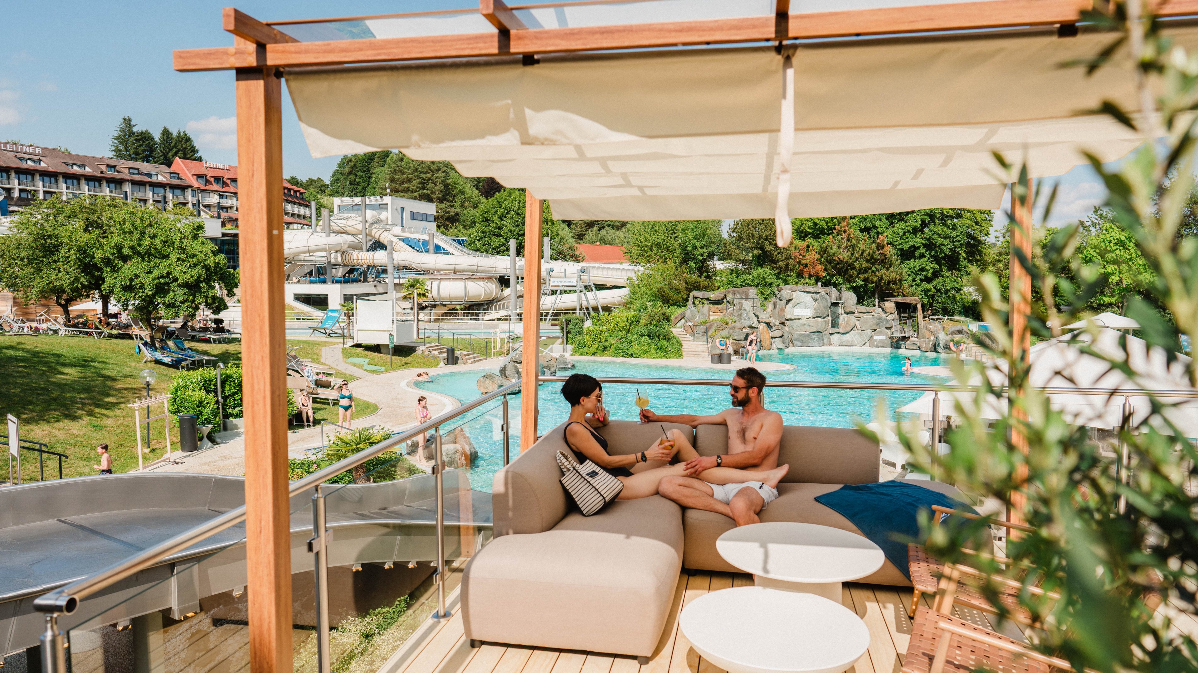 Paar entspannt auf Lounge-Sofa auf Terrasse mit Blick auf Pool und Wasserrutschen im Freibereich der Therme Loipersdorf.
