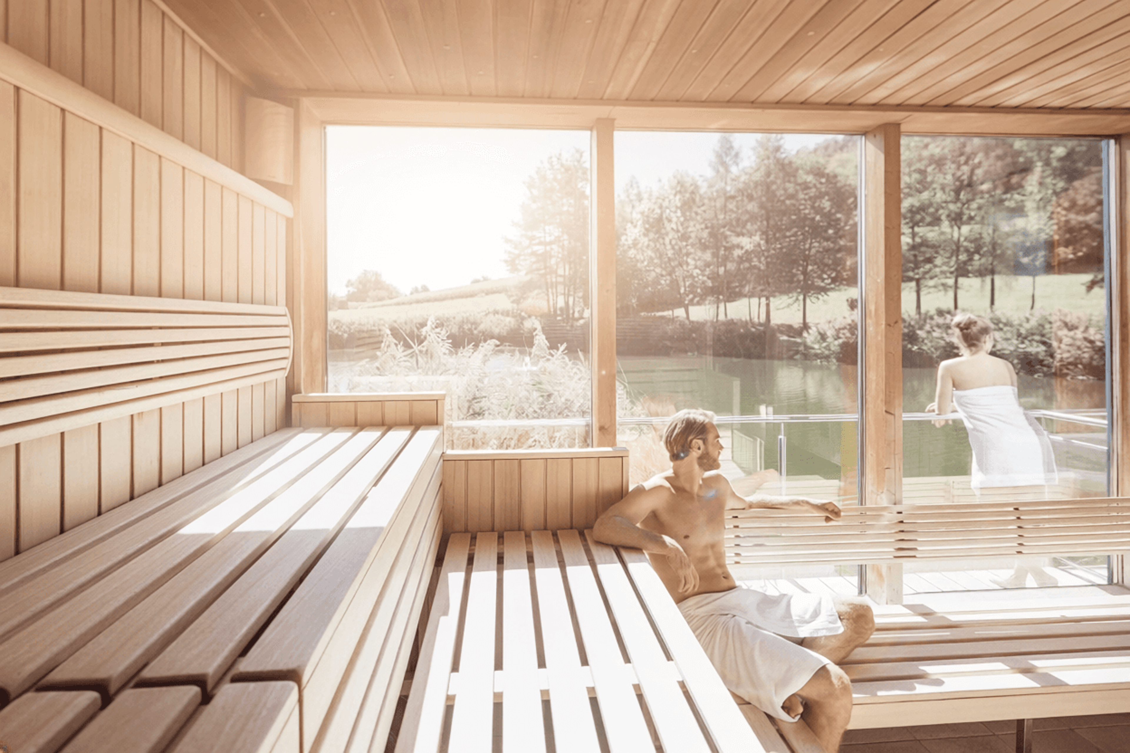 Interior of a sauna with wooden benches and large windows overlooking lush green countryside, with two people in towels relaxing on the benches, one seated and one standing