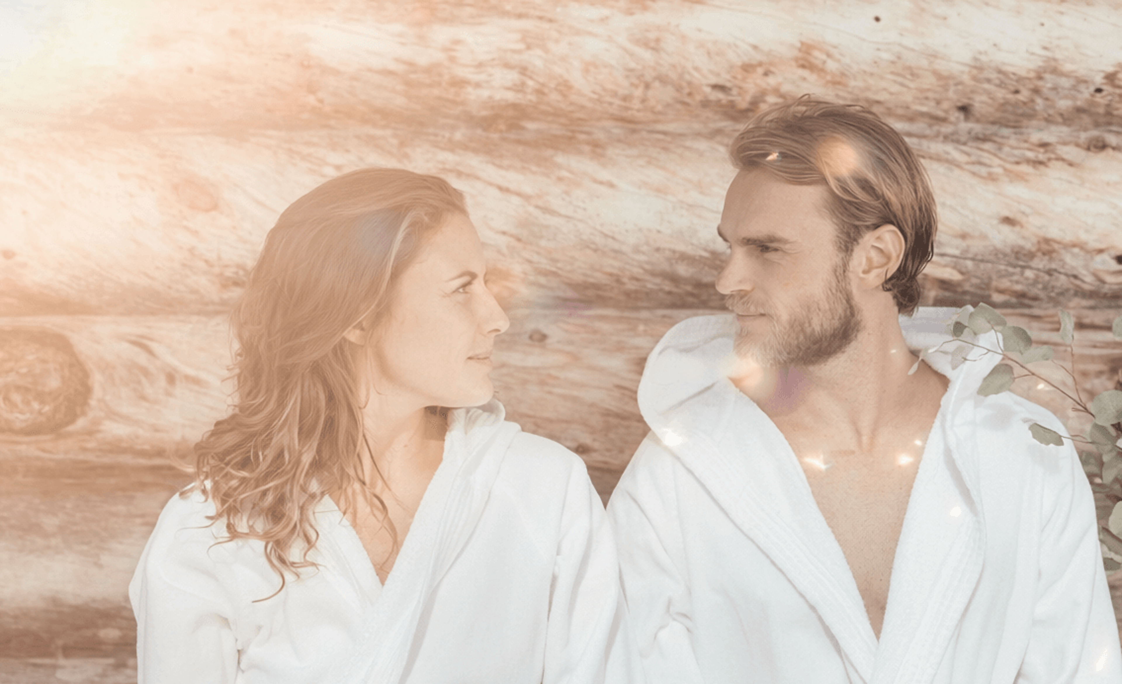 Two people in white bathrobes sit side by side in the sunlight in front of a rocky wall