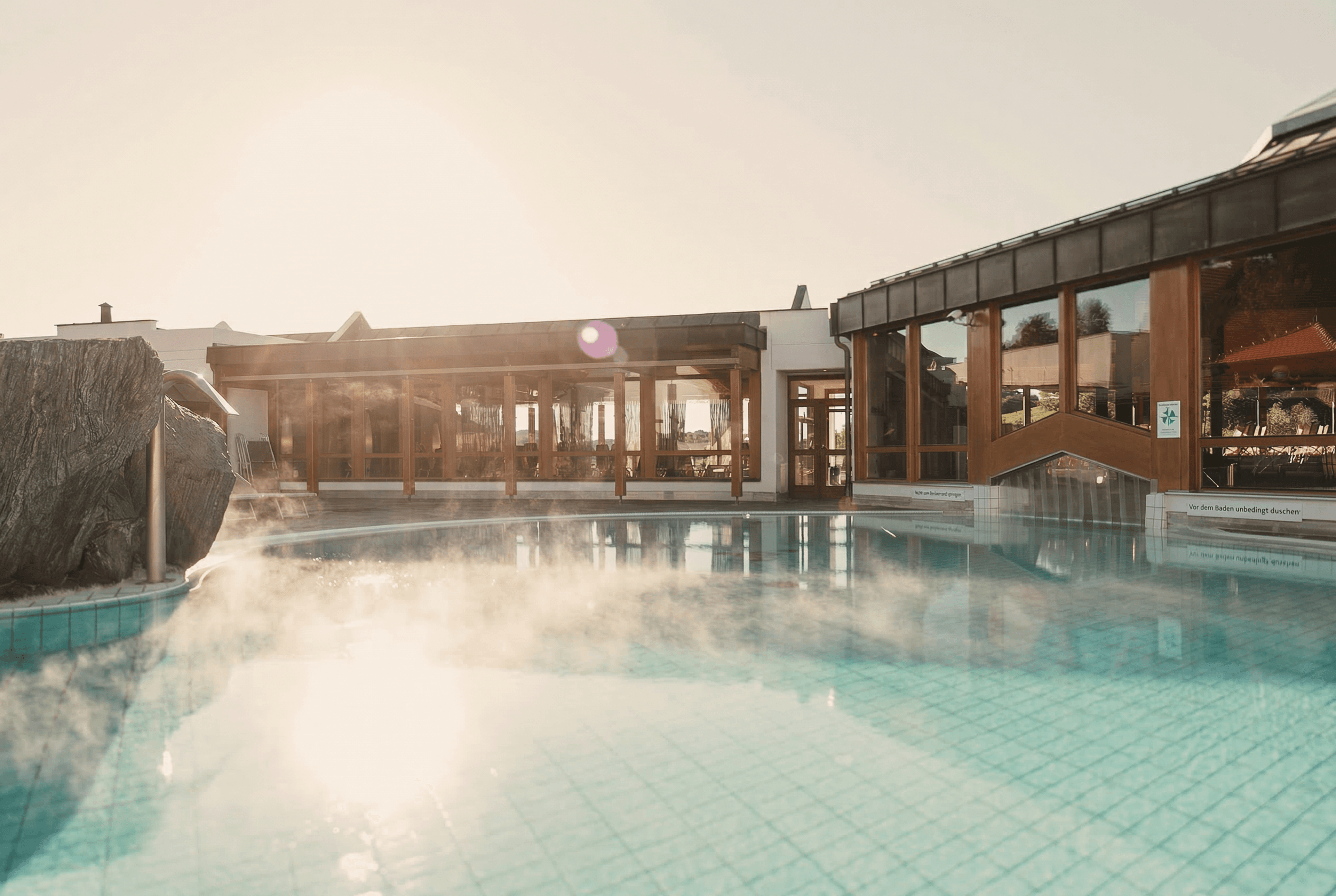 Steaming outdoor pool in front of a building with large glass windows and wooden frames in the sunlight