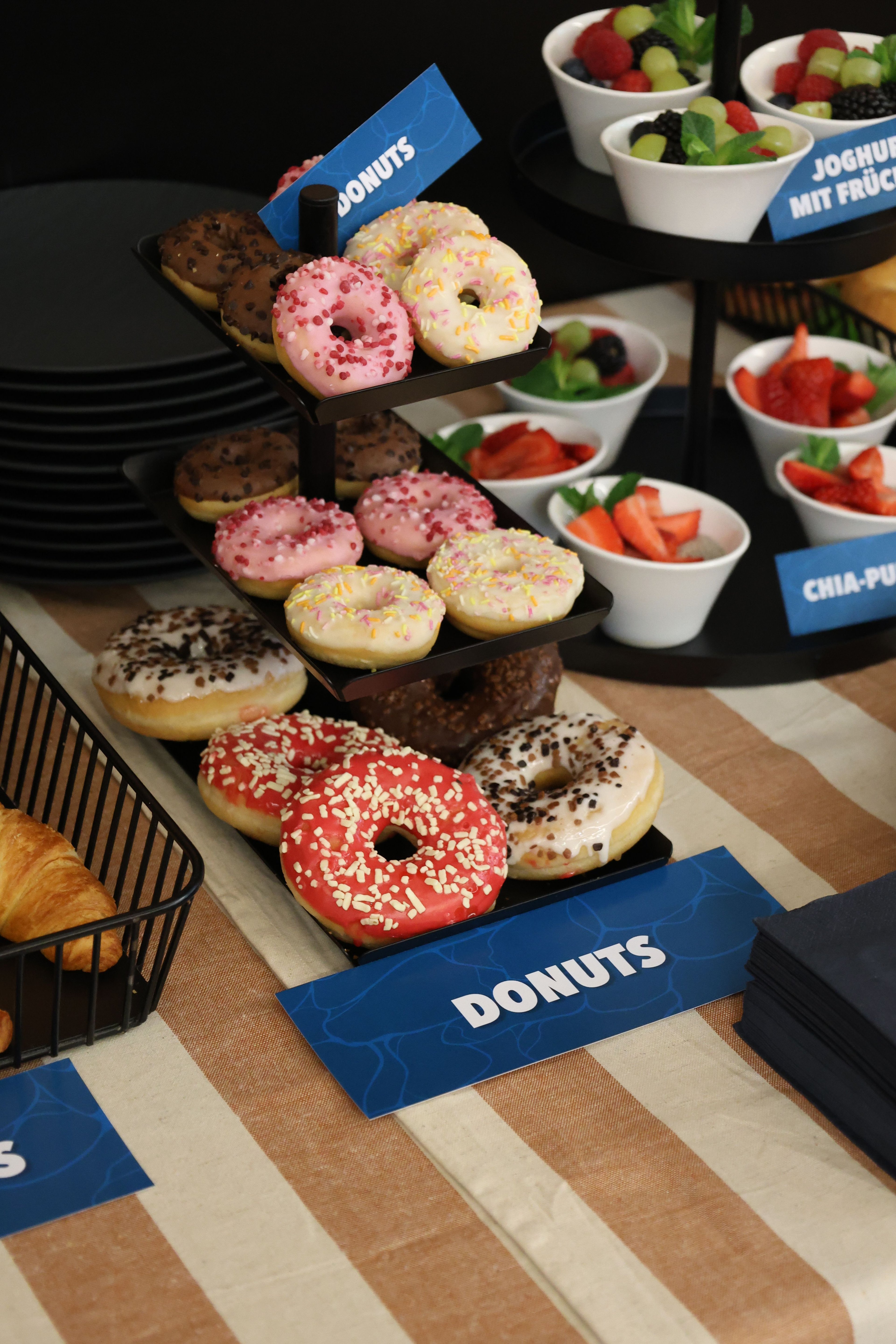 Three-tiered stand with assorted donuts next to bowls of fruit and yogurt on a striped tablecloth.