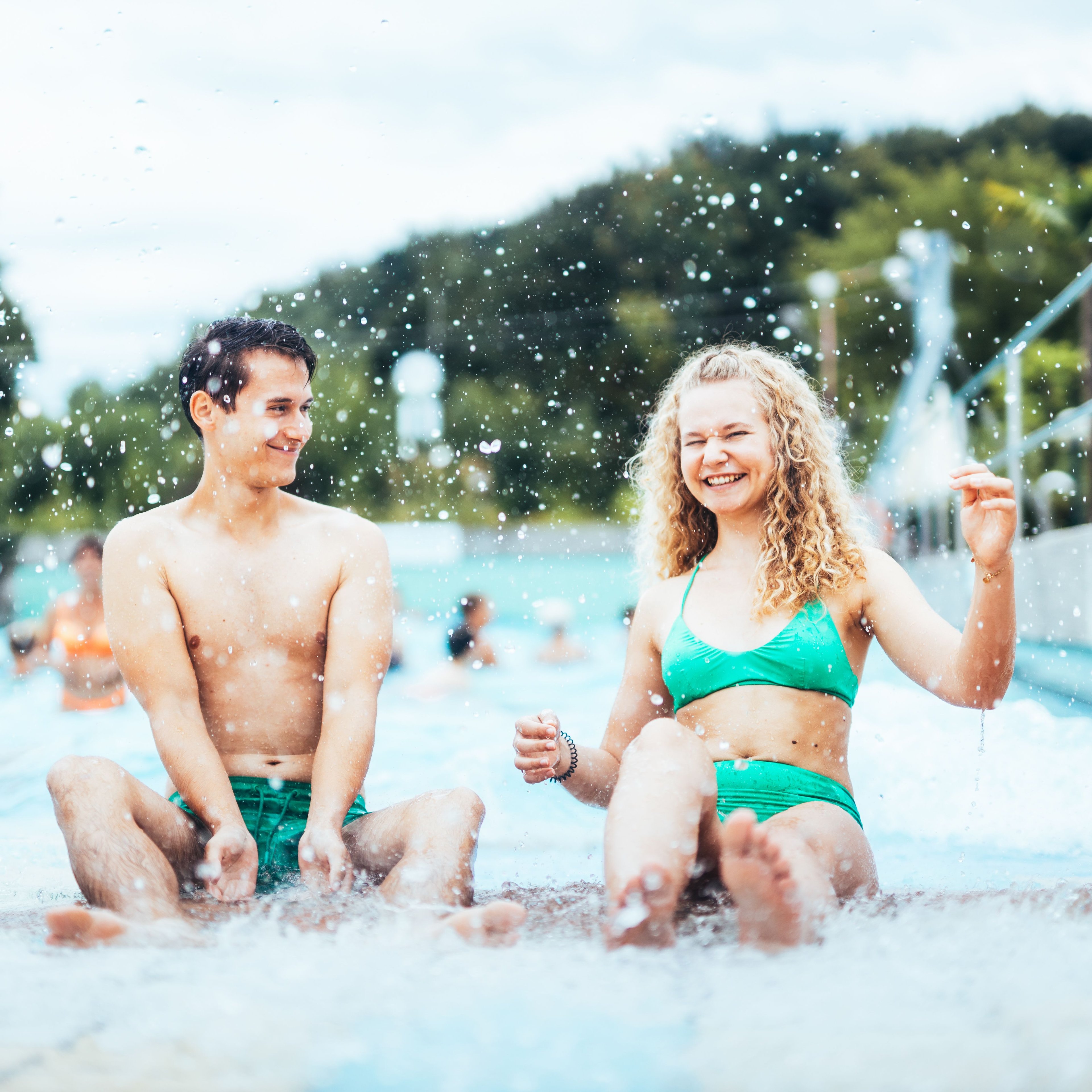 Two young people in swimwear sitting at the edge of a swimming pool splashing water