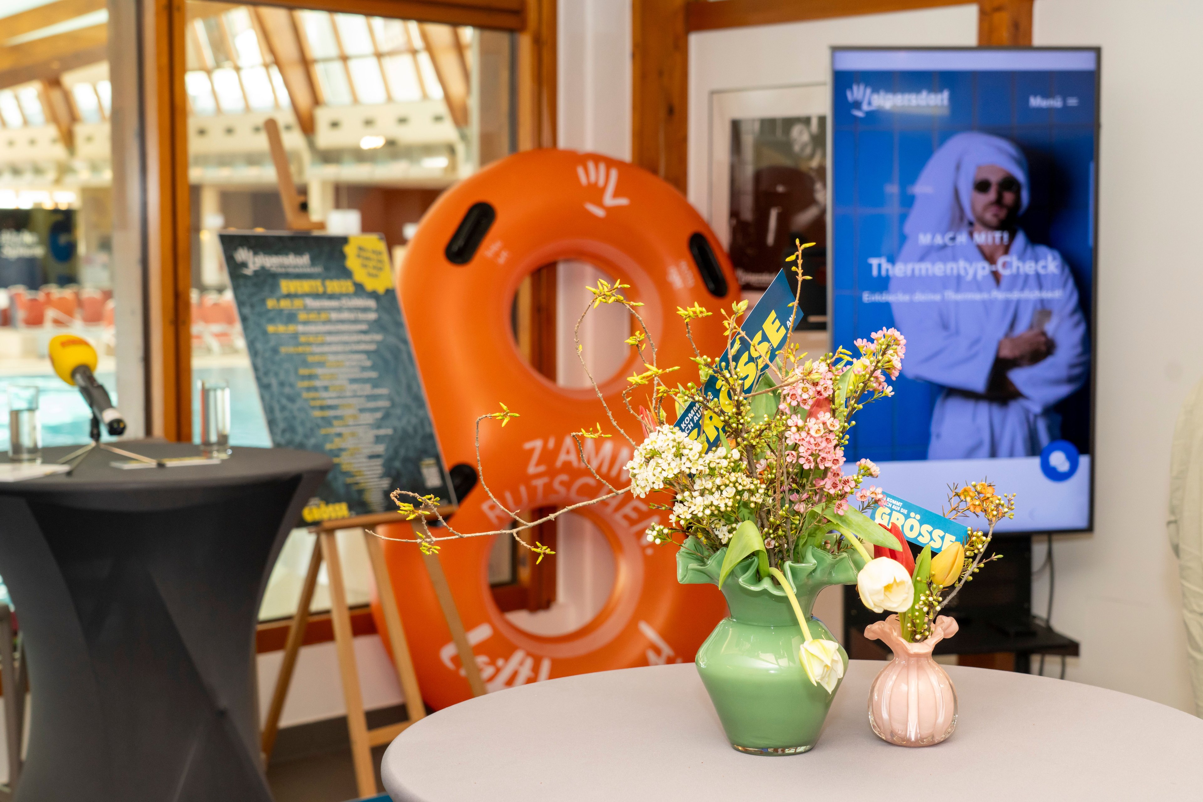 Flower arrangements in two vases on a round table in front of an orange lifebuoy and a screen with blurred image and text.