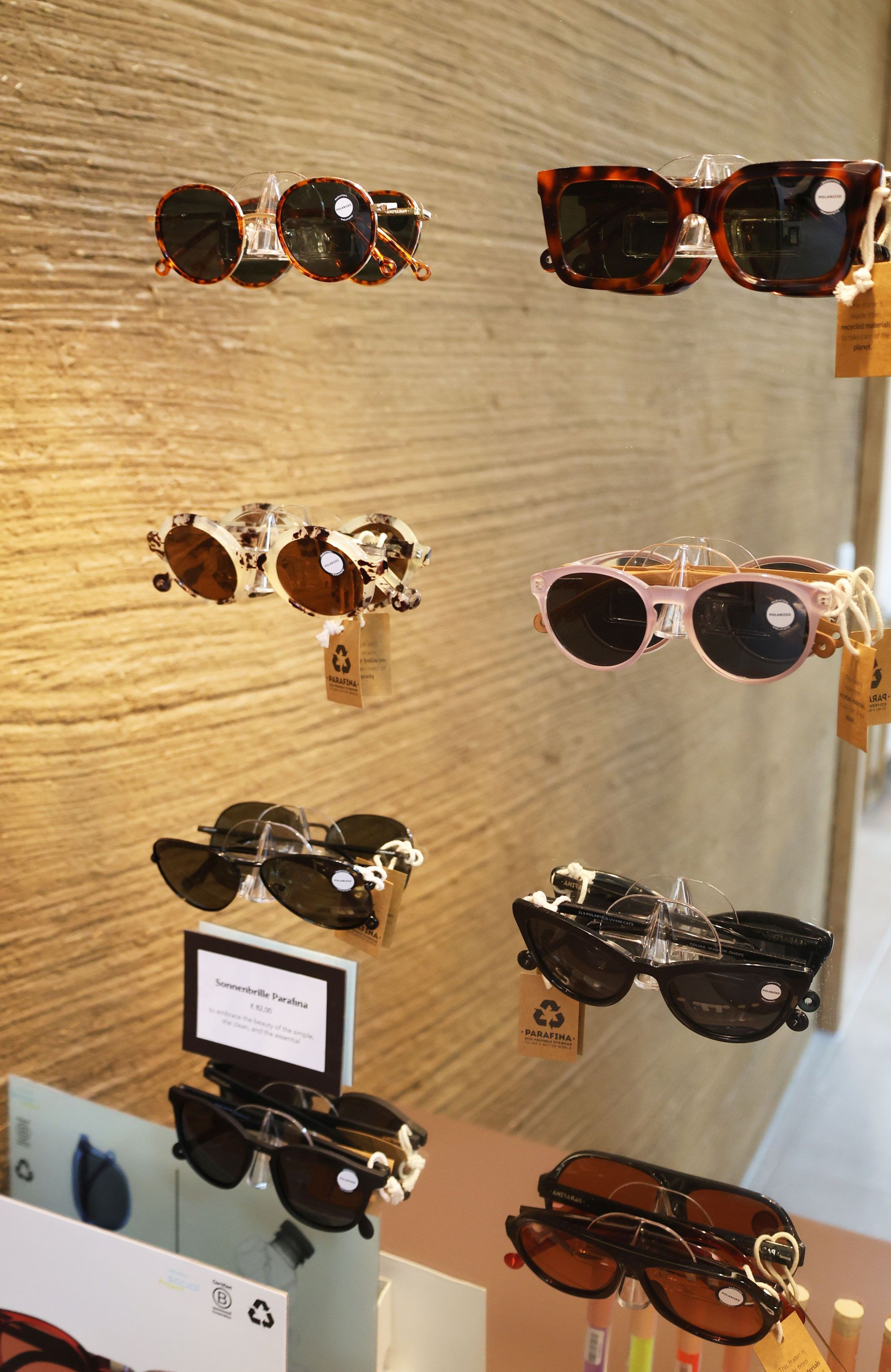Several pairs of sunglasses with different frame colors and shapes, including brown, black, and transparent, on a vertical rack in front of a textured wall.