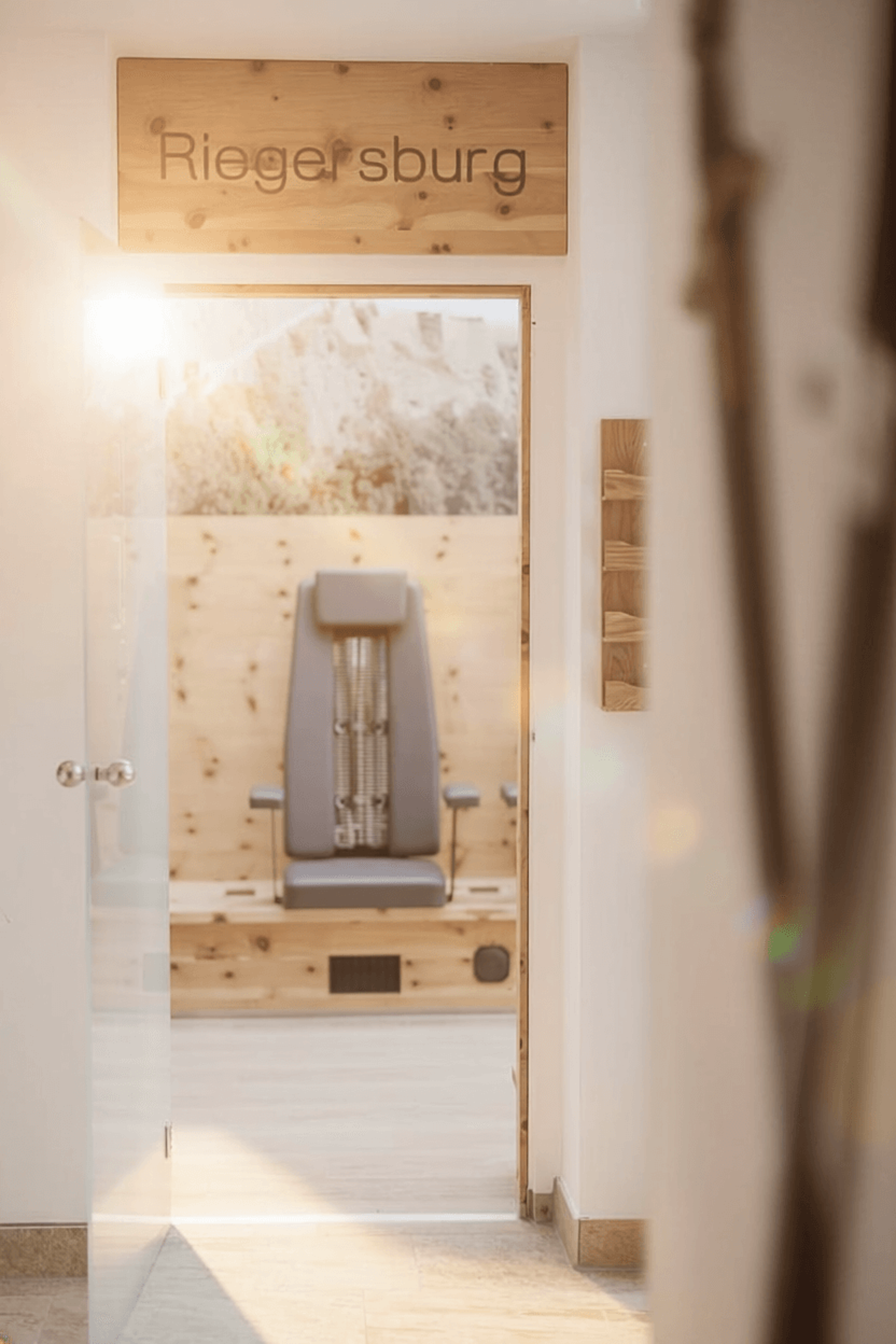 View through an open glass door into a room with a grey massage chair and a wooden sign reading ‘Riegersburg’