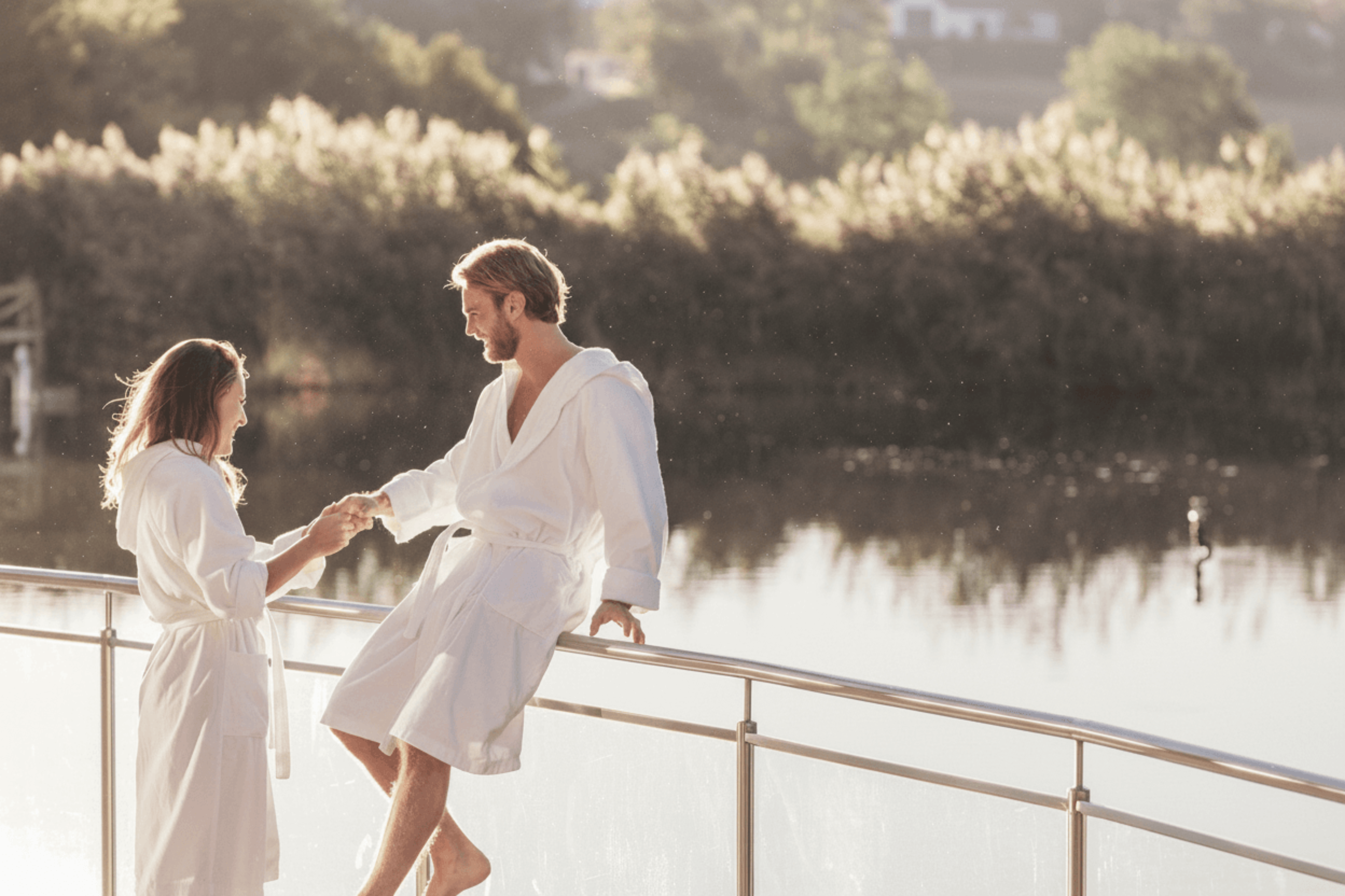 Paar in weißen Bademänteln steht und sitzt an einem Geländer am Wasser mit bewaldetem Hintergrund
