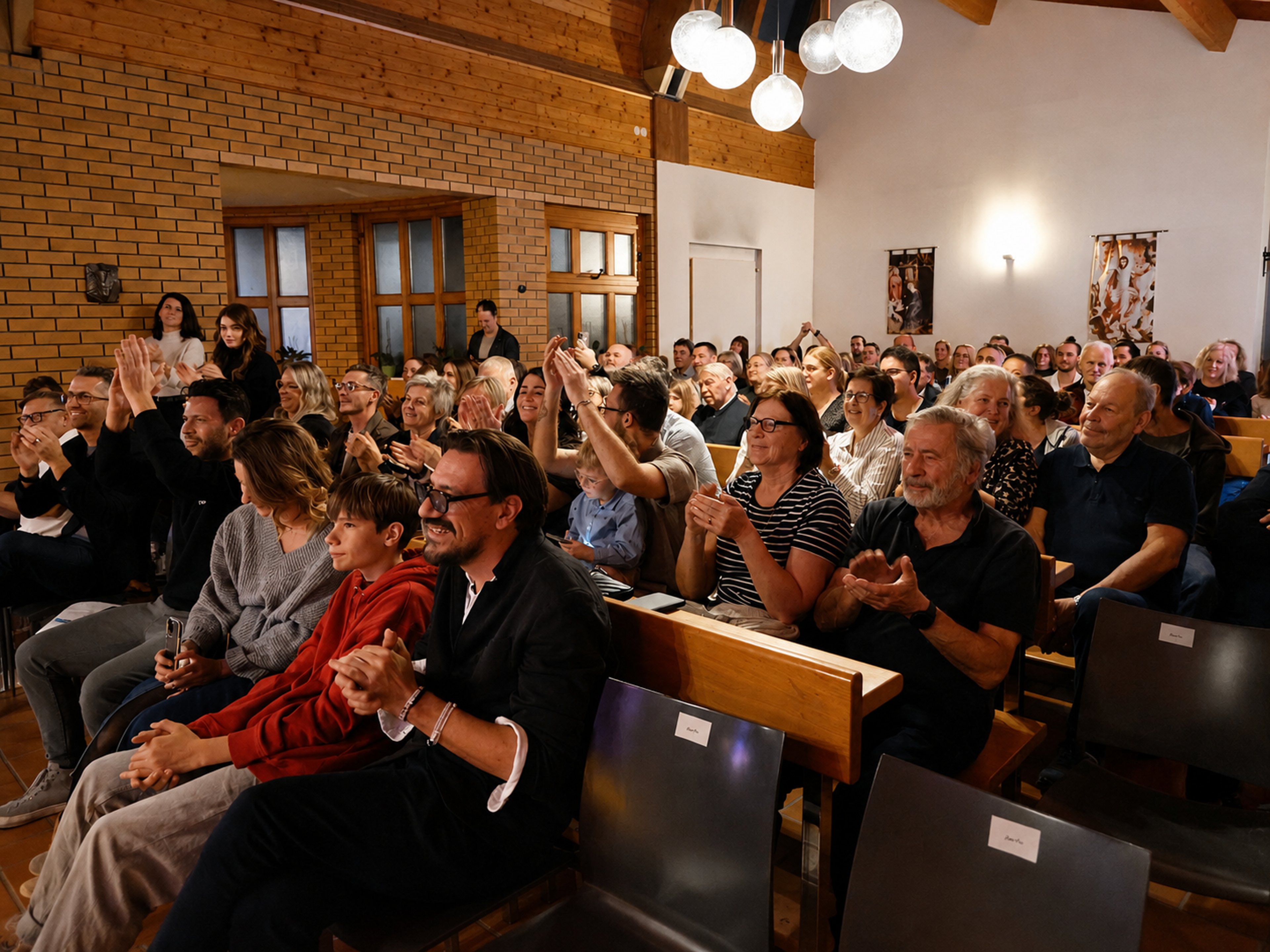 Publikum in einem Saal mit Holzwänden und Decke sitzt auf Bänken und applaudiert