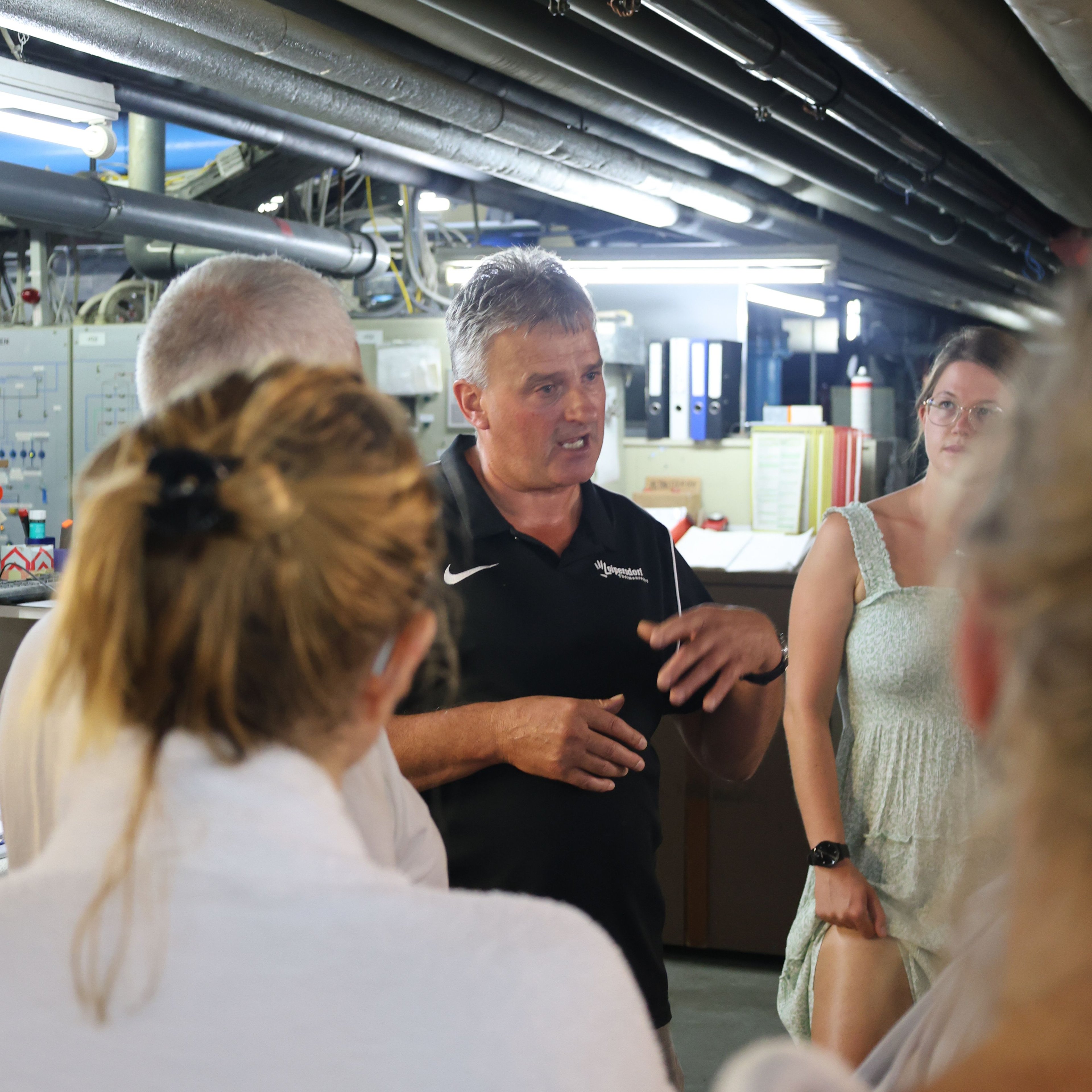 Group of people in a room with visible pipes on the ceiling, one person speaking and gesturing
