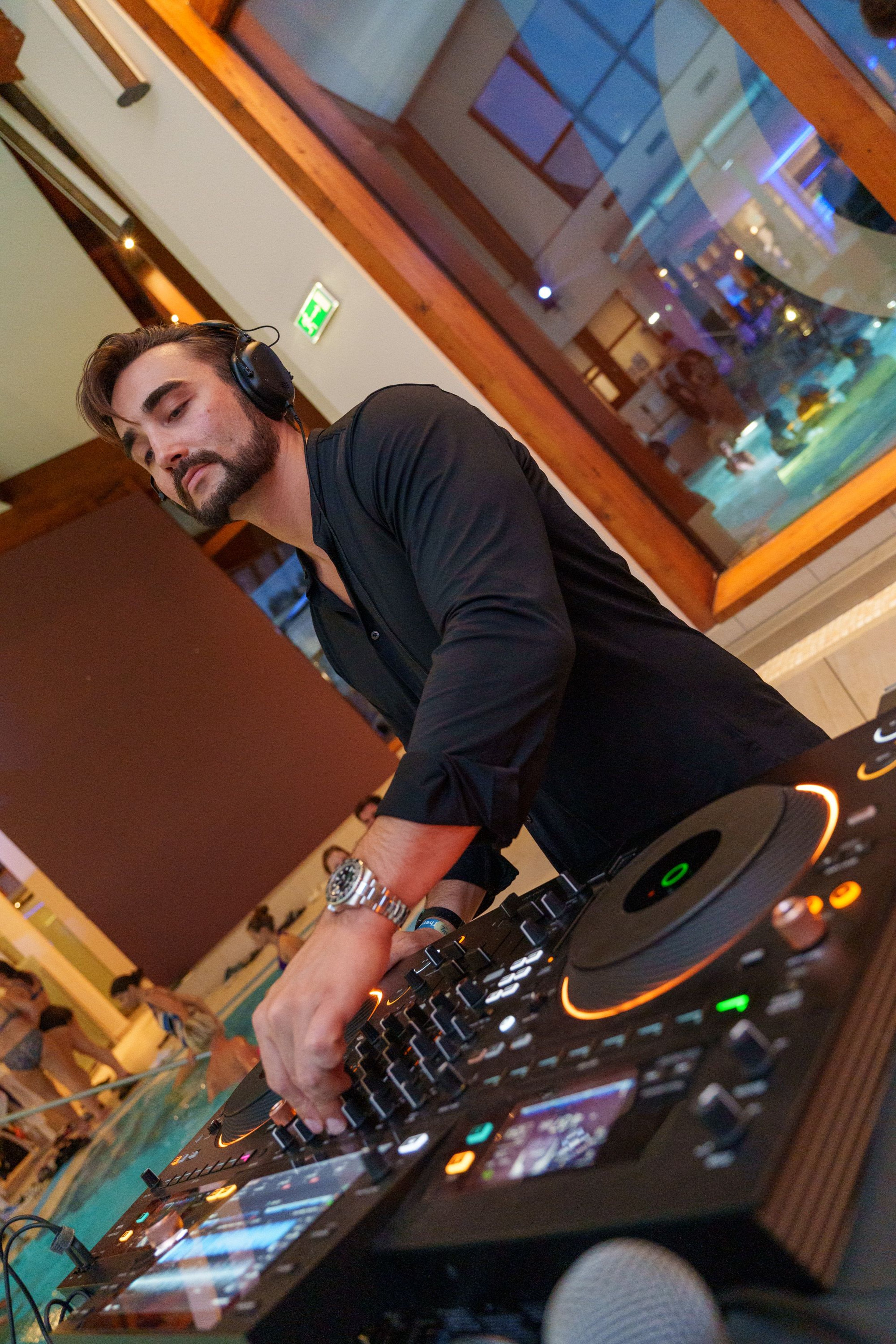 Mann mit Kopfhörern bedient DJ-Mischpult in Innenraum mit Pool im Hintergrund