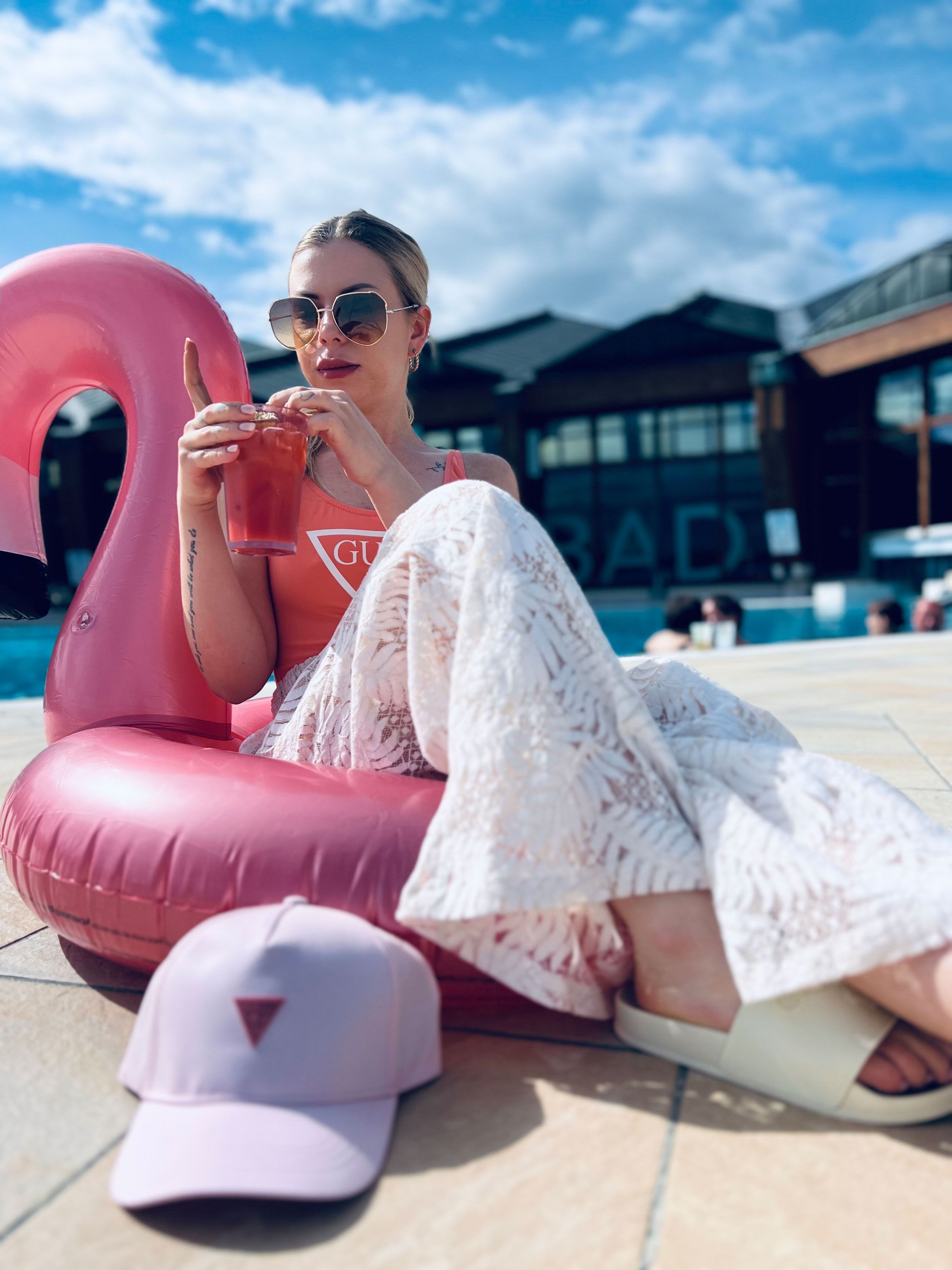 Person sitting on an inflatable pink flamingo, wearing patterned white pants and white sandals, holding a drink in their hand, with a light pink cap with a red triangle logo in front of them.
