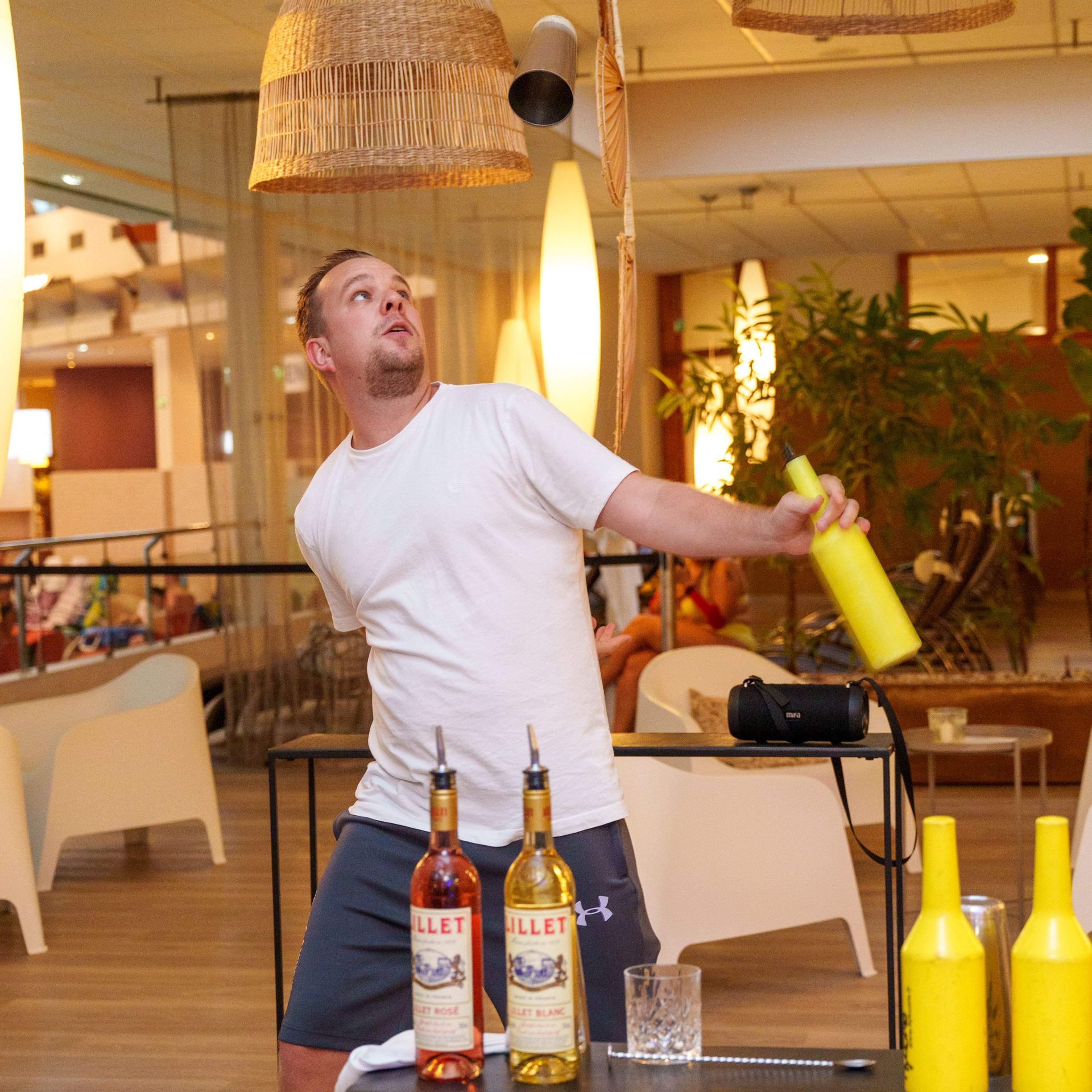 A show bartender throws a yellow bottle in the air in a modern bar with wicker pendant lamps and seating in the background