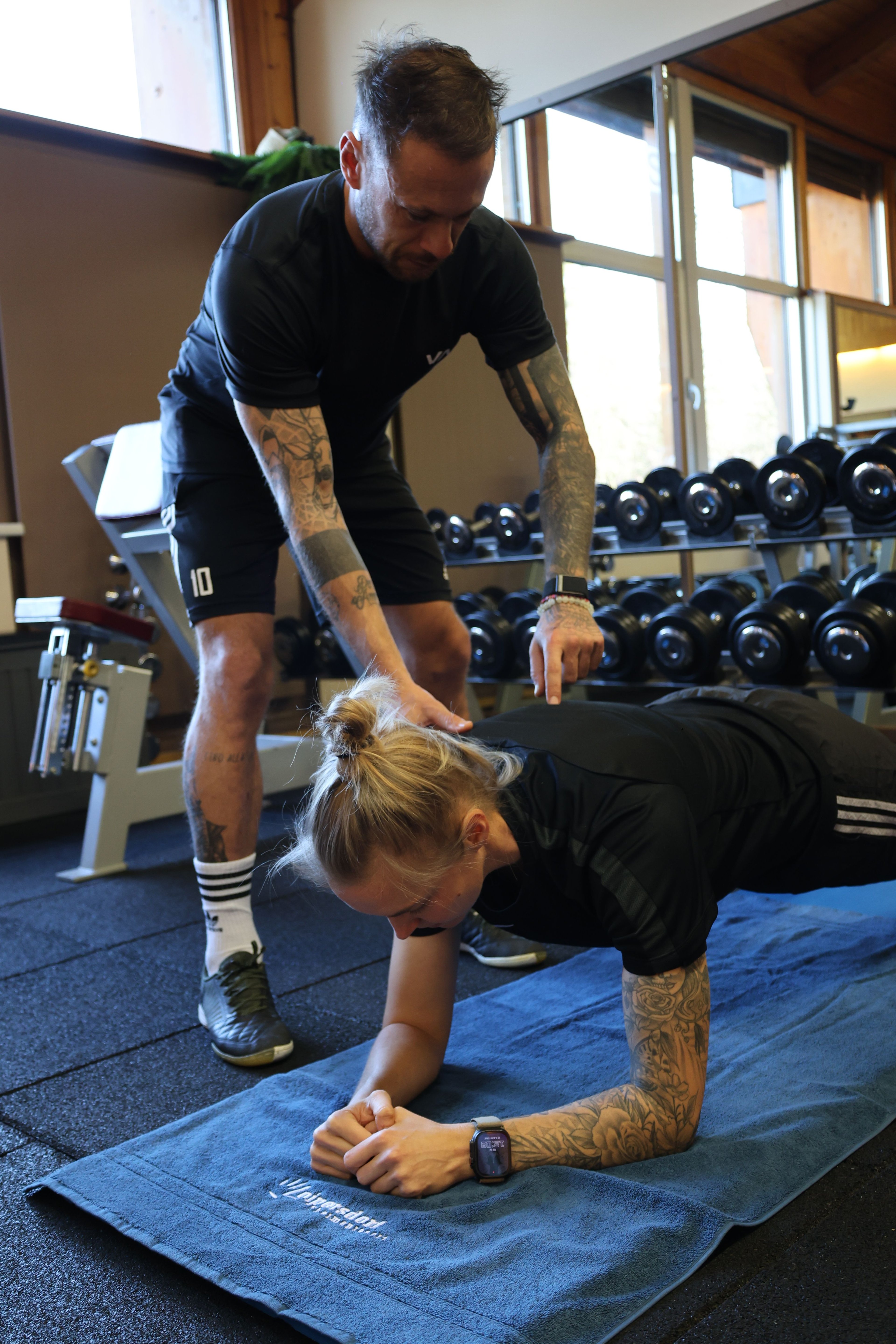 Man with tattoos and sportswear assisting woman with tattoos doing a plank exercise on a blue mat in a gym