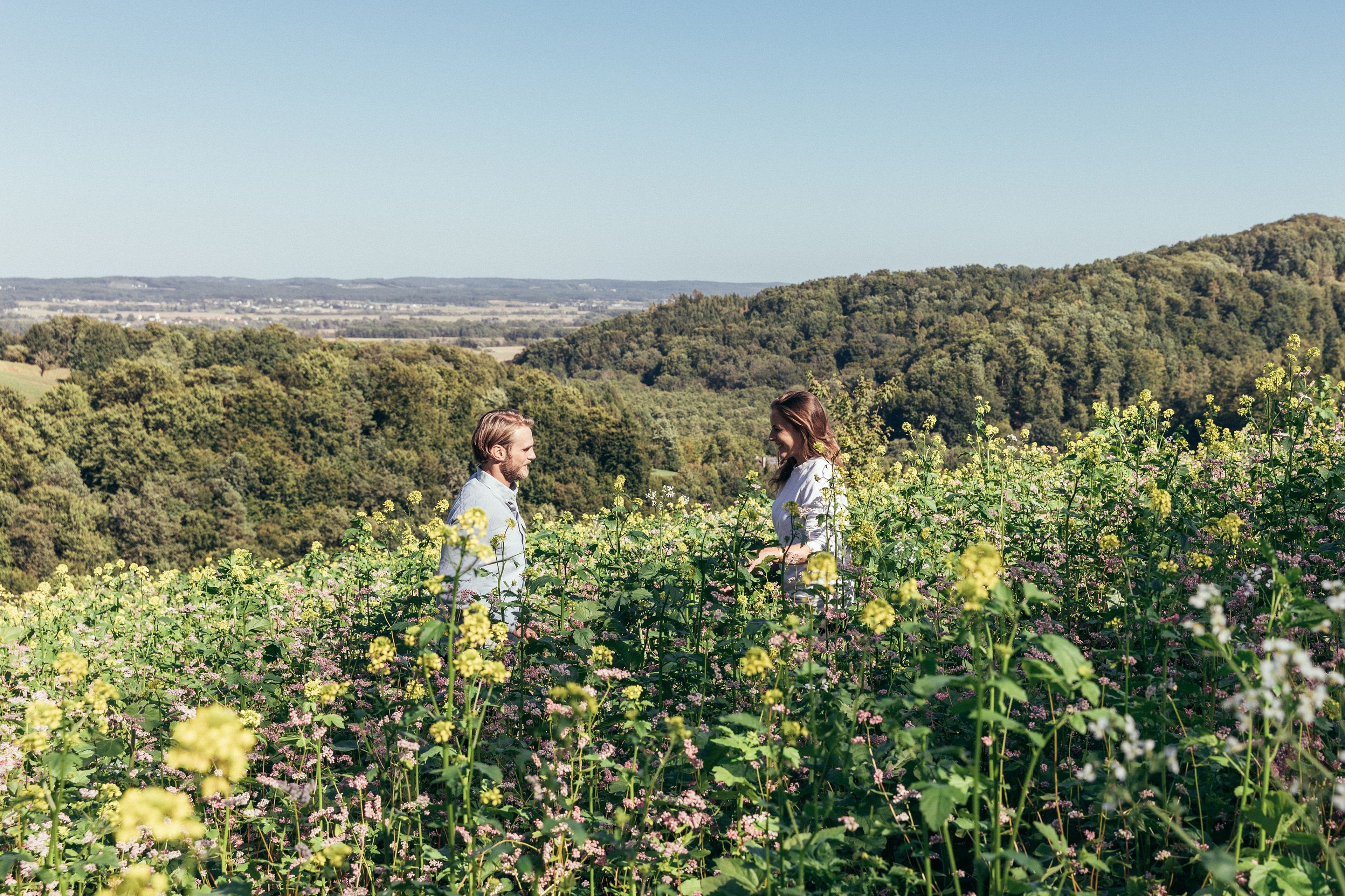 Couple standing in the meadow and the landscape around Bad Loipersdorf can be seen in the background.