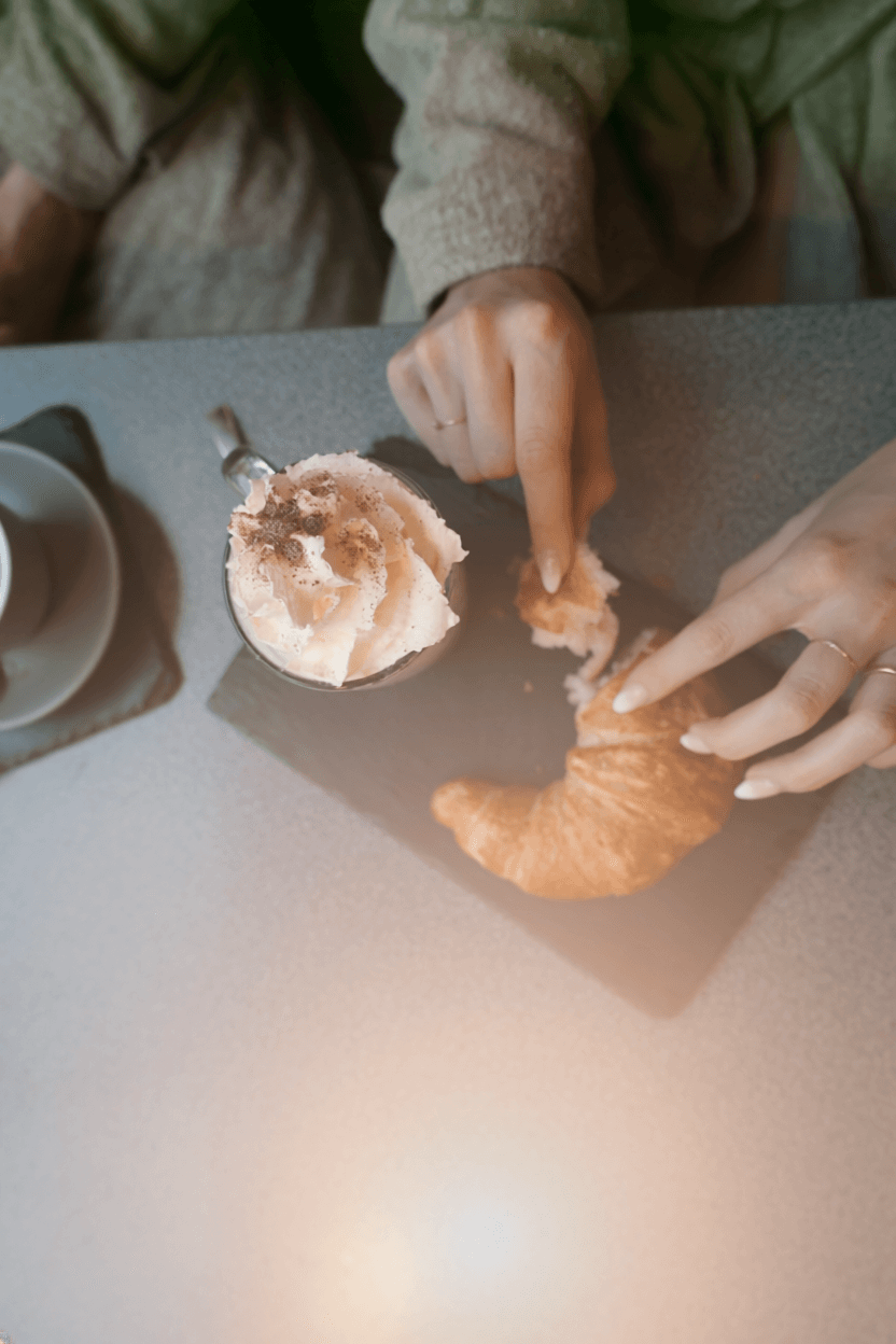 Two hands reach for a croissant next to a cup of coffee topped with whipped cream on a table