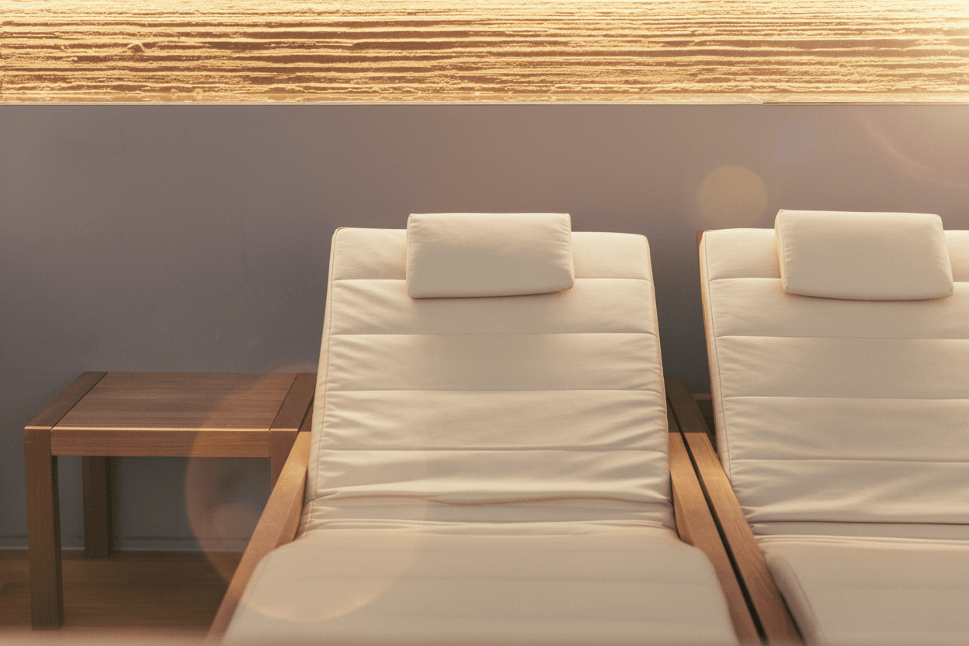 Zwei gepolsterte Liegen mit Kopfstützen neben einem kleinen Holztisch vor einer grauen Wand
