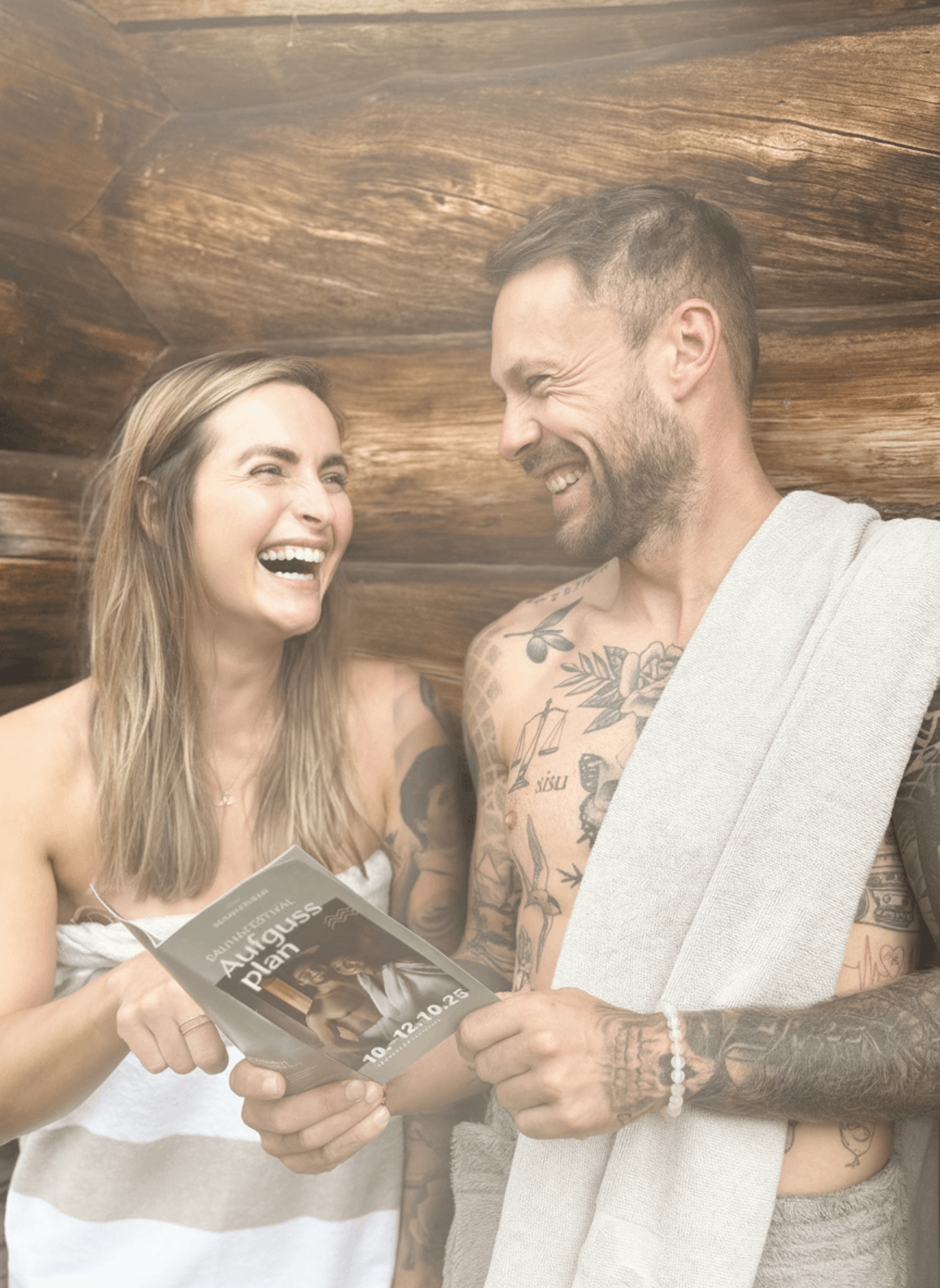 Zwei tätowierte Personen in Saunatüchern lesen gemeinsam einen Plan in einer Holzsauna