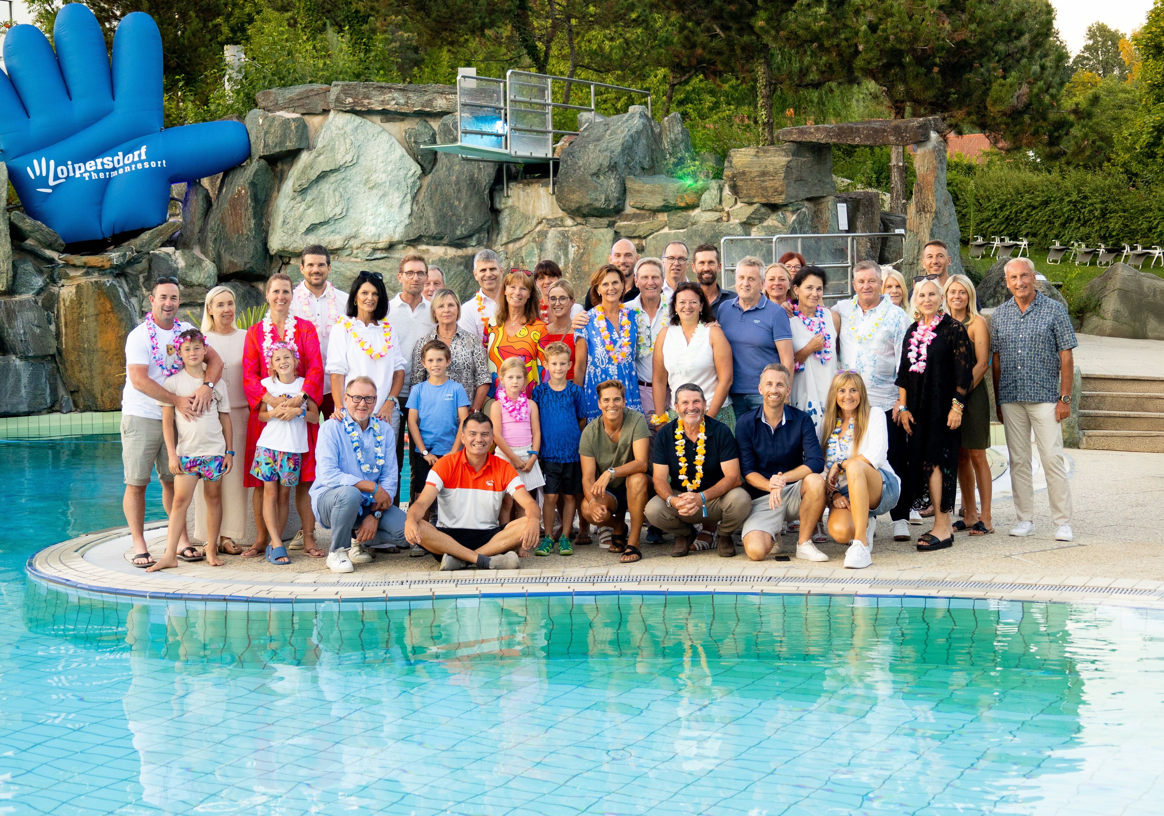 Large group of people of various ages wearing flower leis standing and sitting at the edge of a swimming pool in front of rocks and trees
