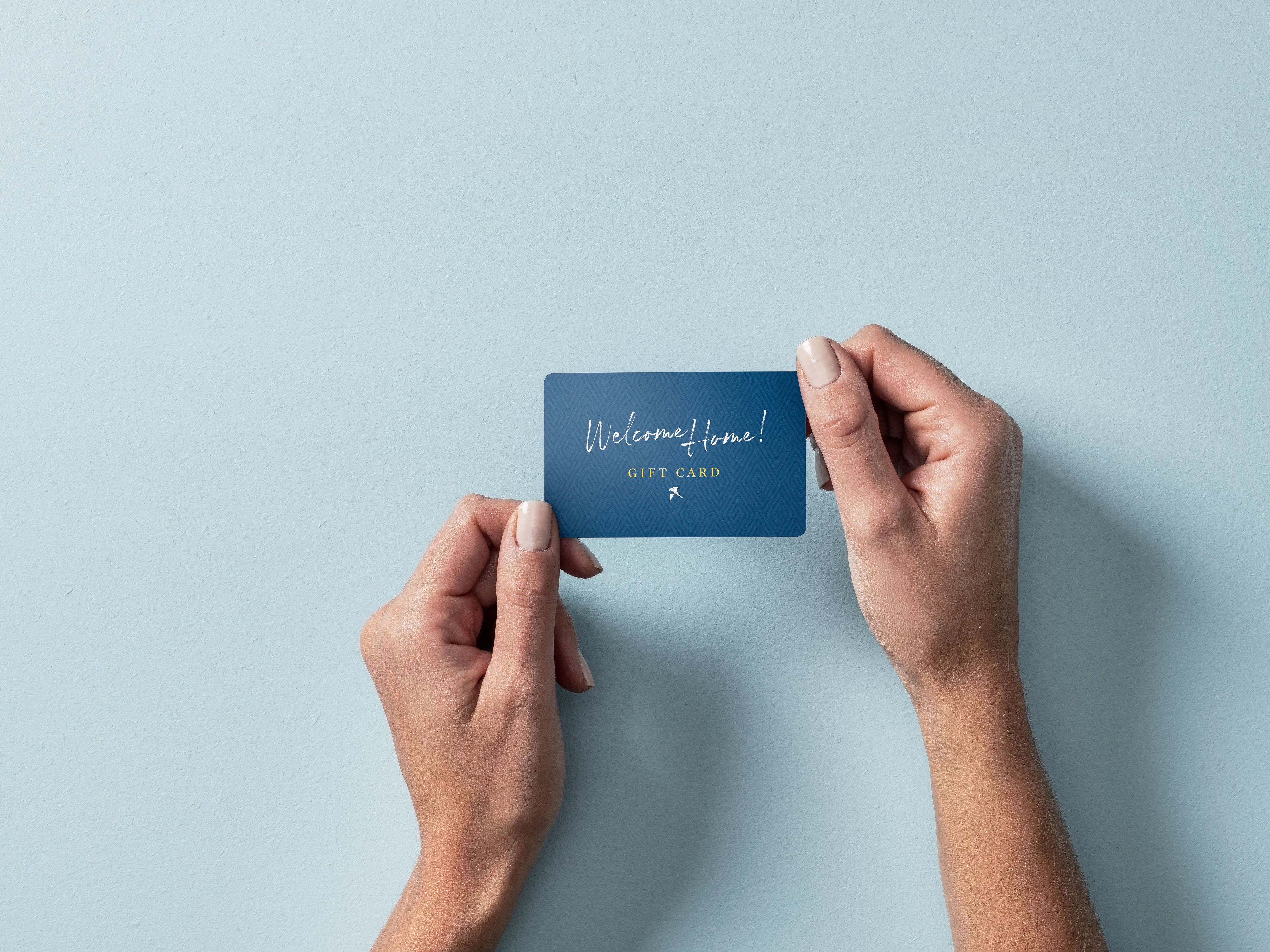 Zwei Hände halten eine blaue Geschenkkarte mit der Aufschrift 'Welcome Home! GIFT CARD' vor hellblauem Hintergrund