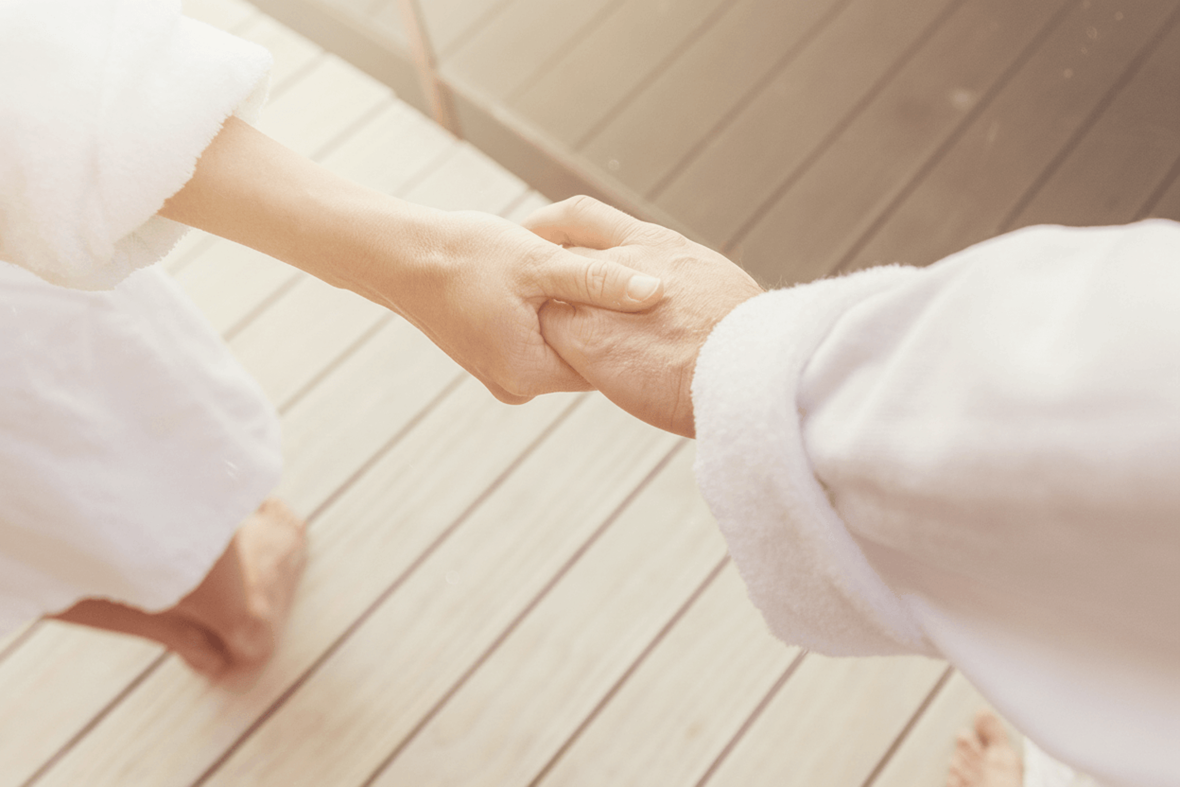 Zwei Personen in Bademänteln halten sich an den Händen stehend auf einem Holzfußboden