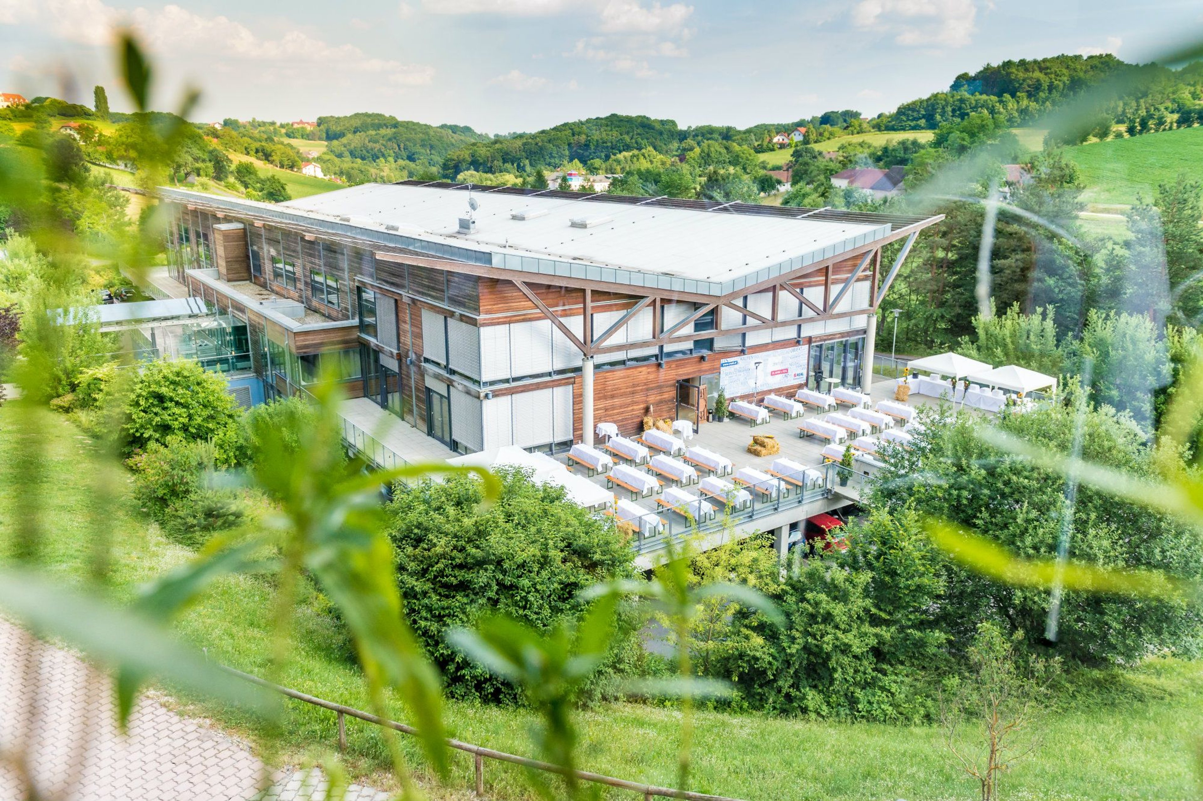 Modern two-story wooden building with large terrace and multiple tables in a green hilly landscape