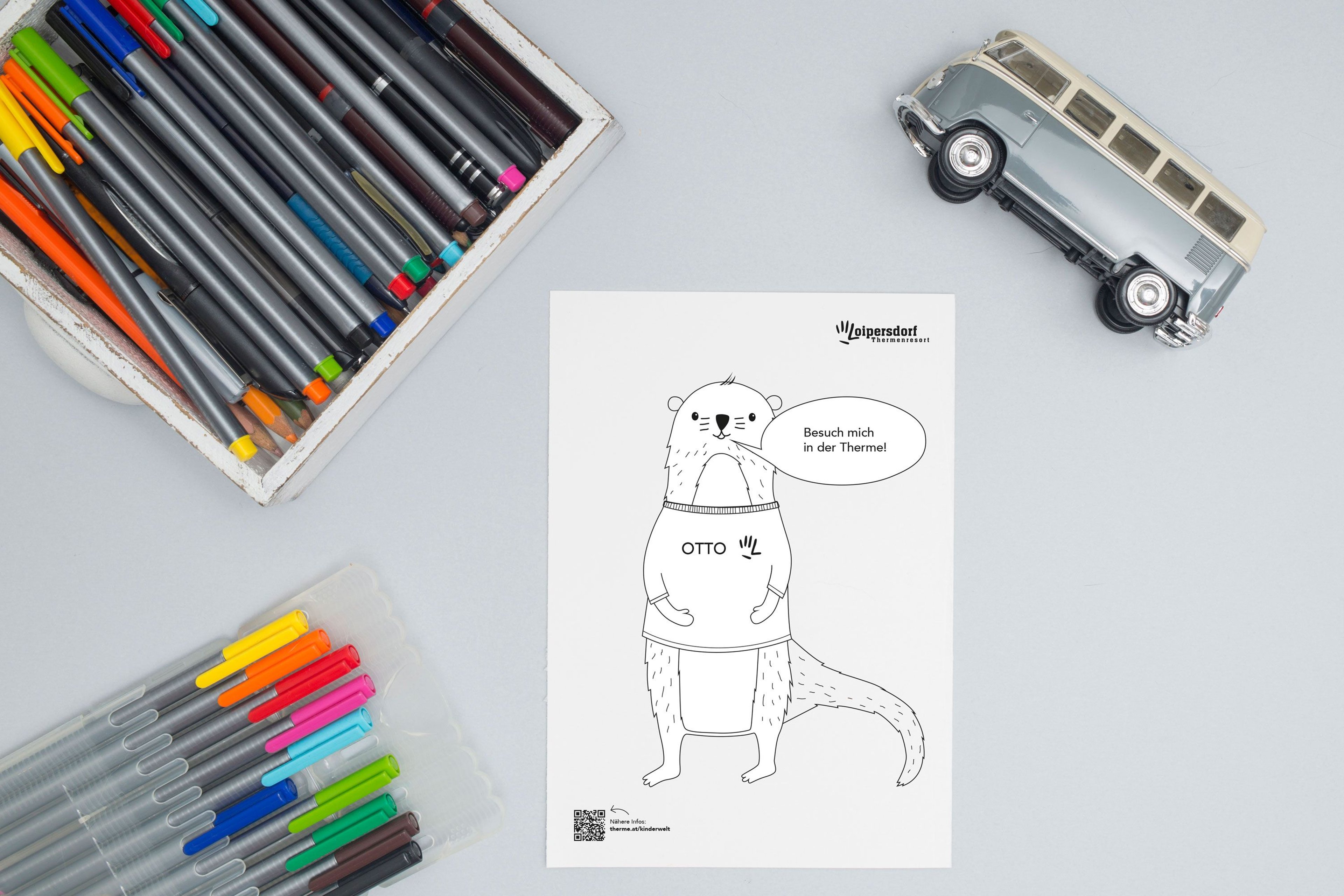 Top view of a table with a coloring picture of Otto, the spa mascot. Next to it are colorful pencils and a small toy bus.