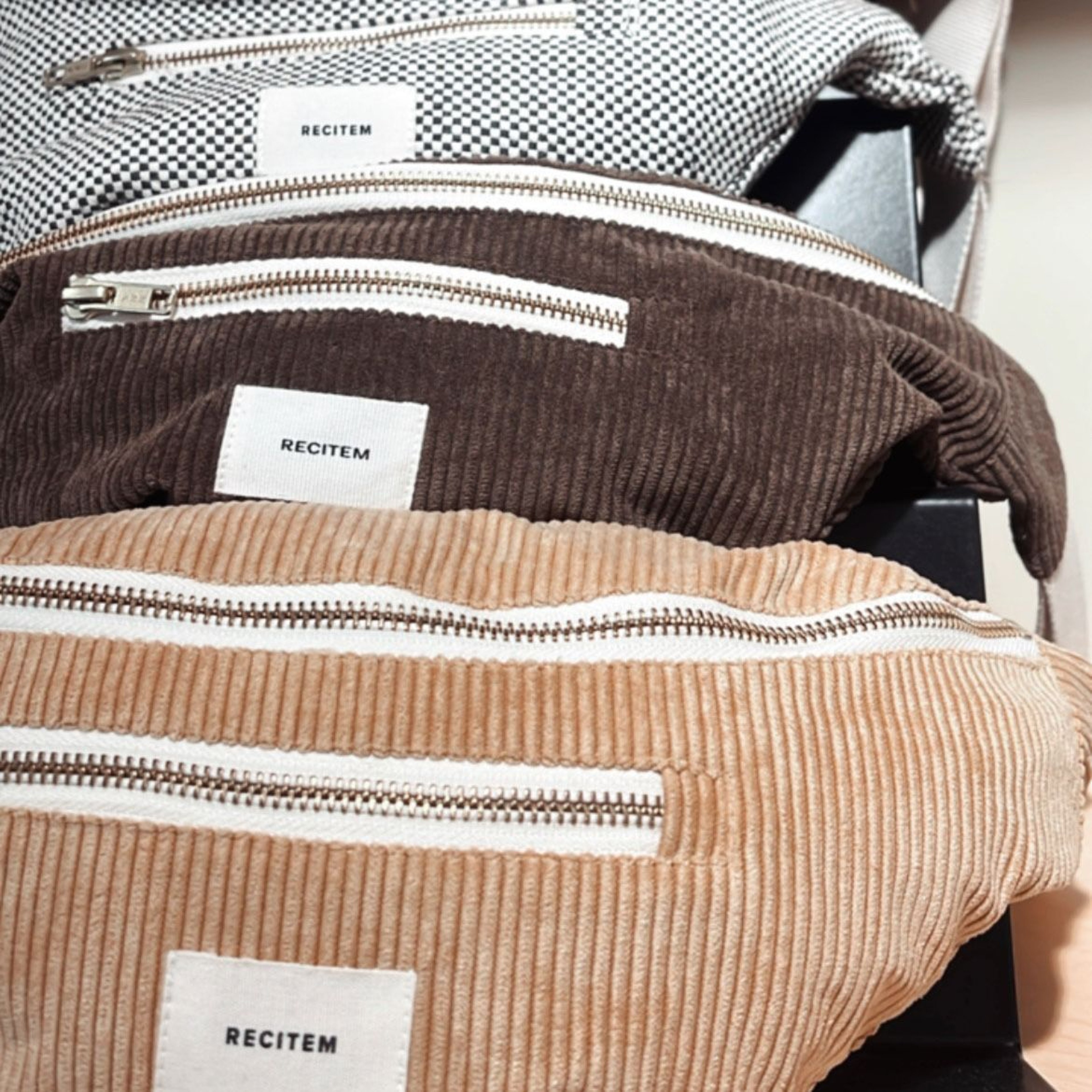 Three corduroy bum bags in beige, brown, and black and white with white zippers and labels with lettering.