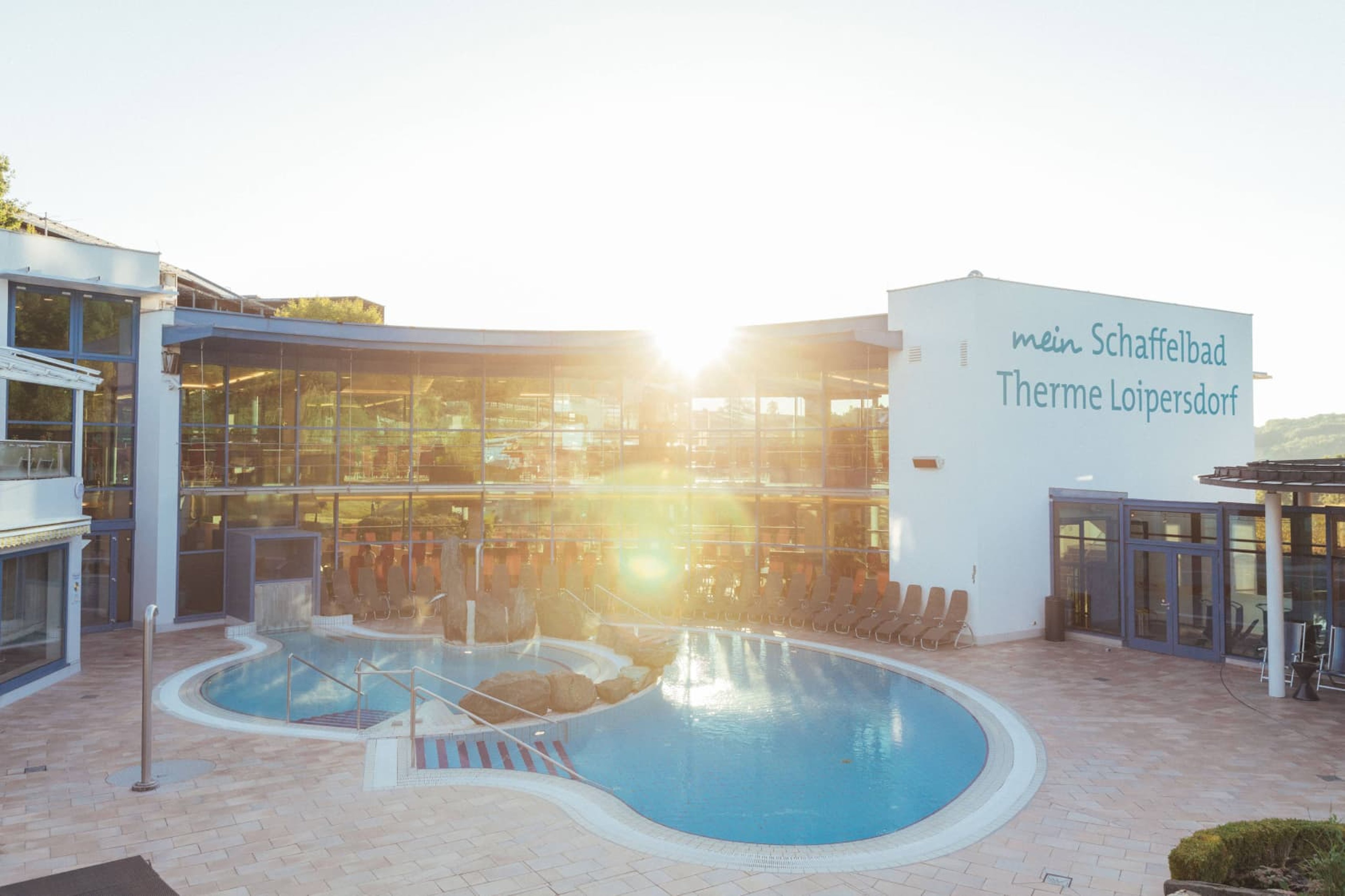 Ausblick auf das Thermalbecken im Schaffelbad. Im Vordergrund das Becken mit dem blauen Thermalwasser und im Hintergrund die Architektur des Schaffelbades mit Sonnenstrahlen. 