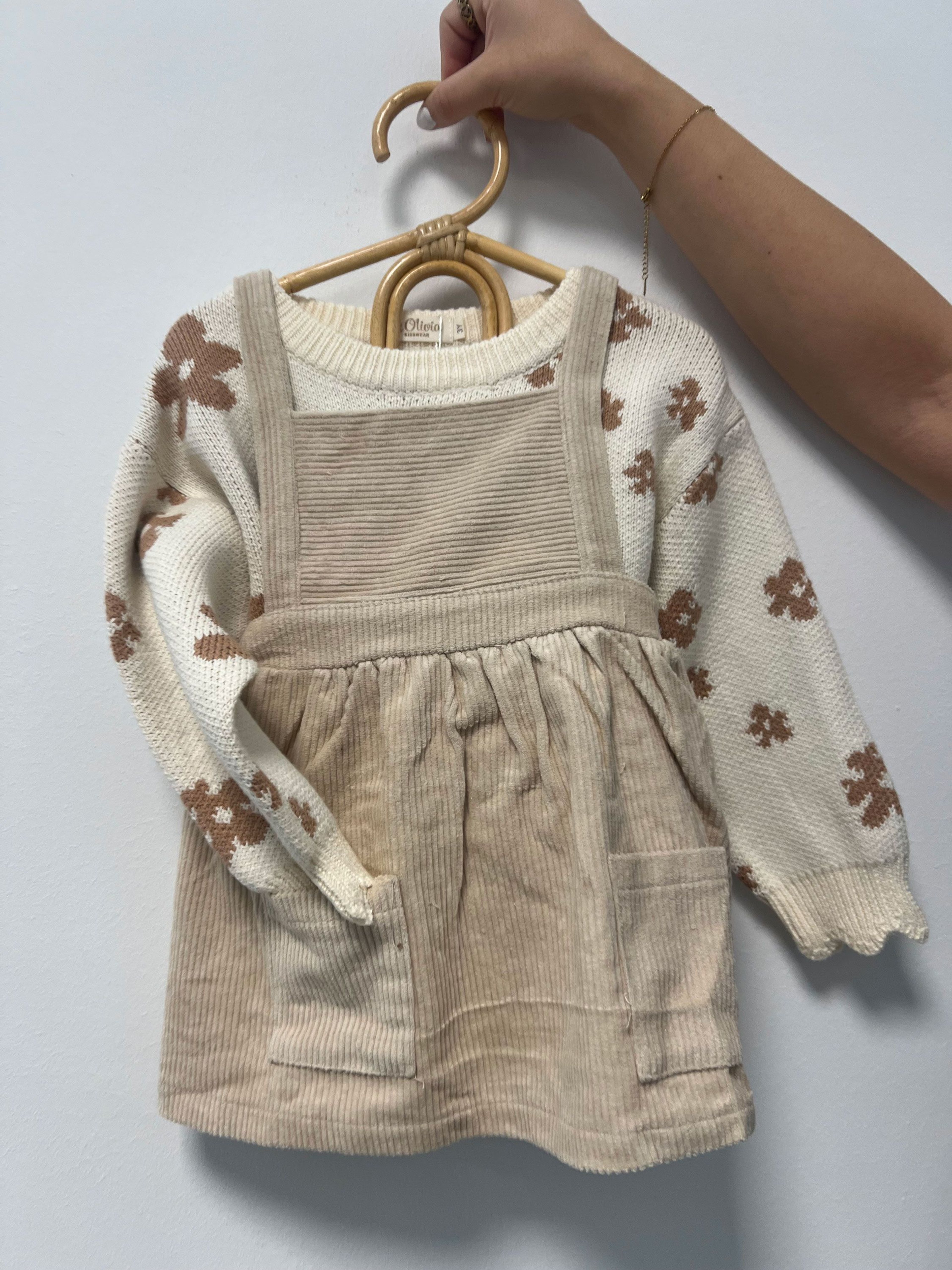 Beige corduroy dress with pockets over a cream-colored sweater with brown floral pattern, held on a wooden hanger.