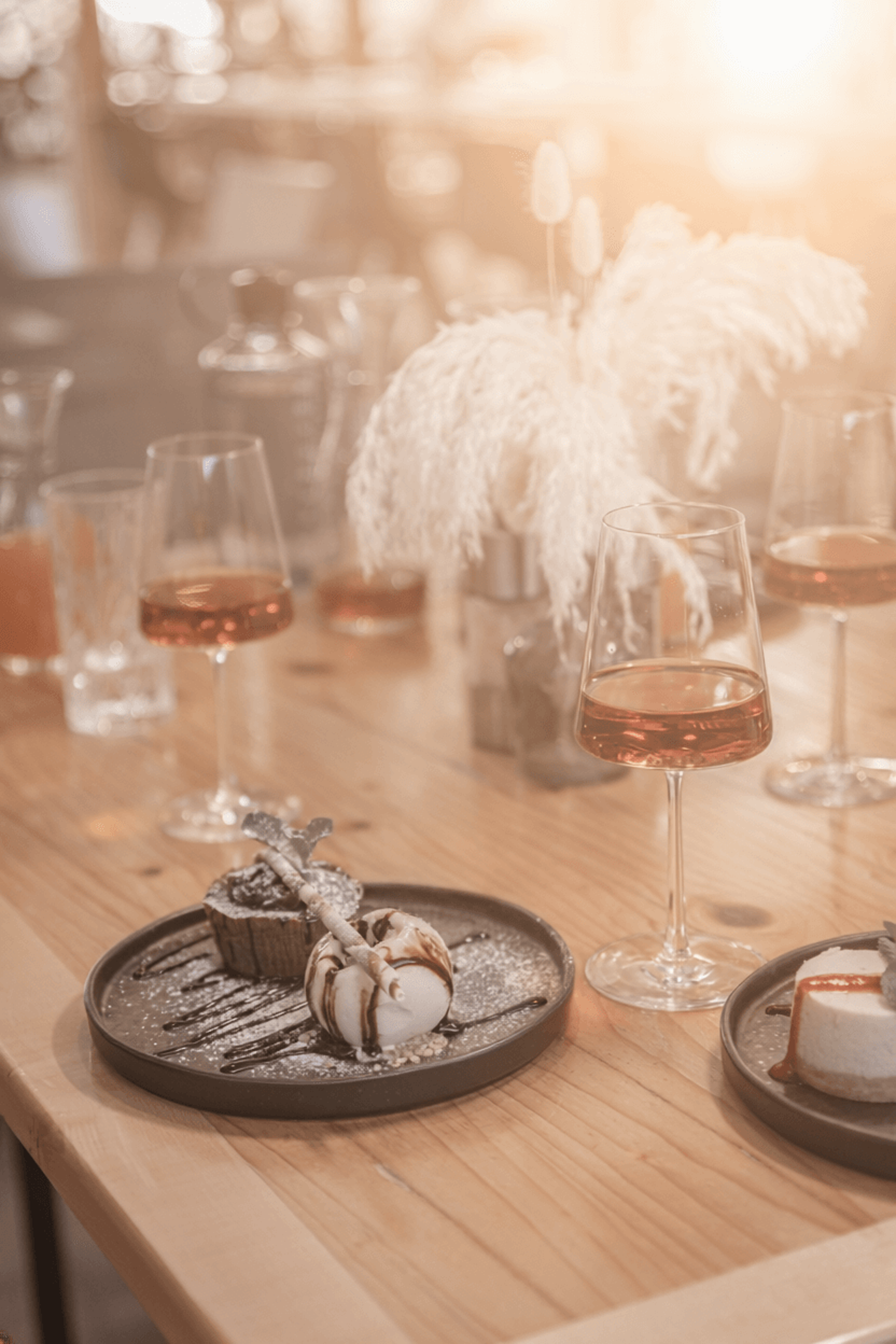Dessert plate with chocolate cake and vanilla ice cream on a wooden table, next to glasses of rosé-colored drinks and decorative white feathers