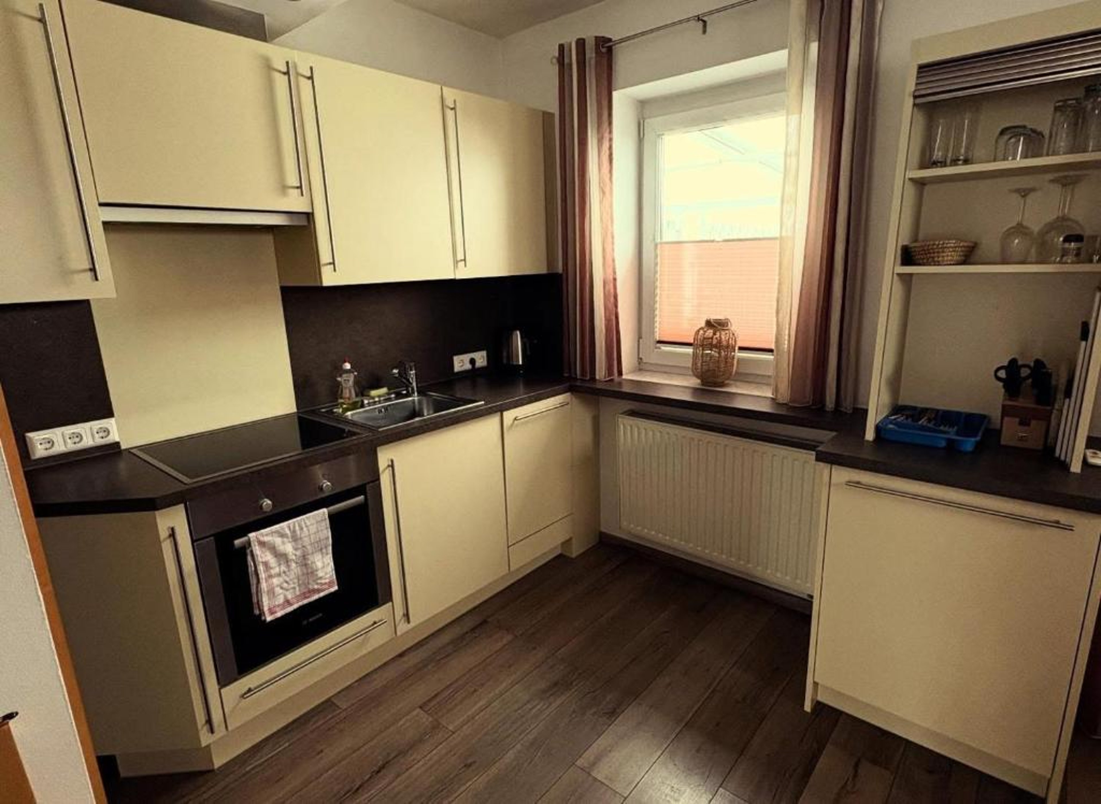 Small kitchen with light cabinets, black countertop, built-in oven, and window with curtains