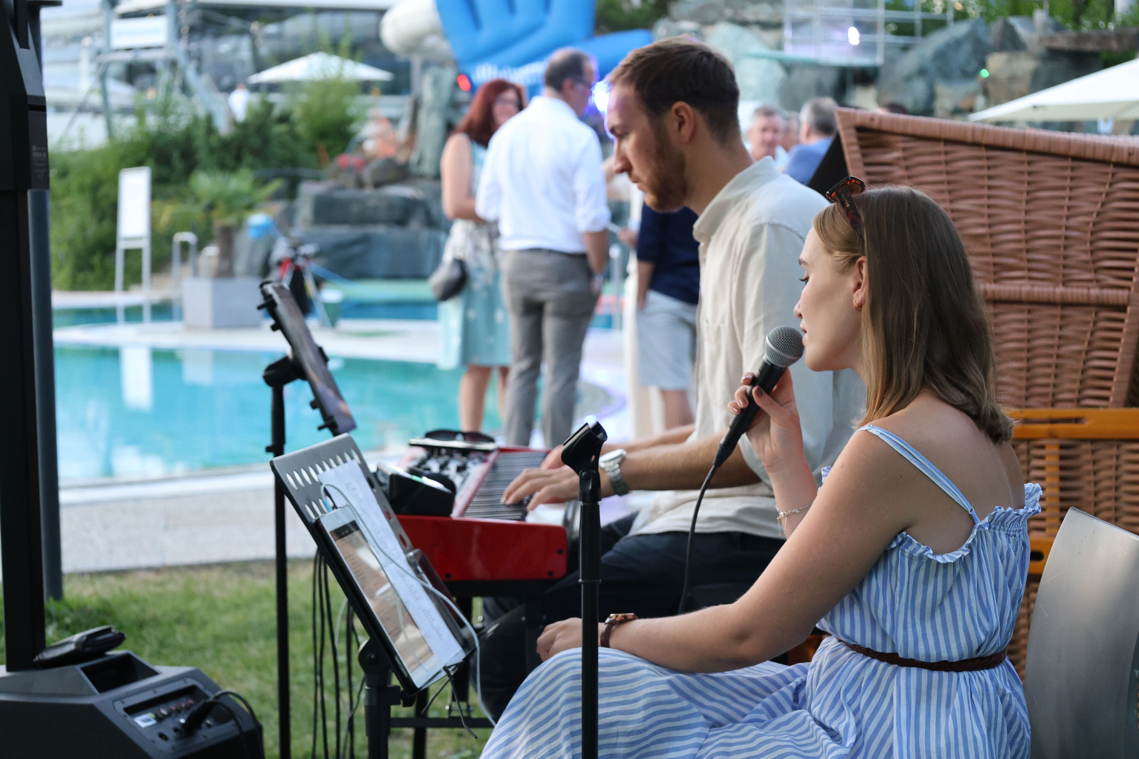 Woman in striped dress holding a microphone sitting in front of a man playing a keyboard outdoors at an event with several people in the background
