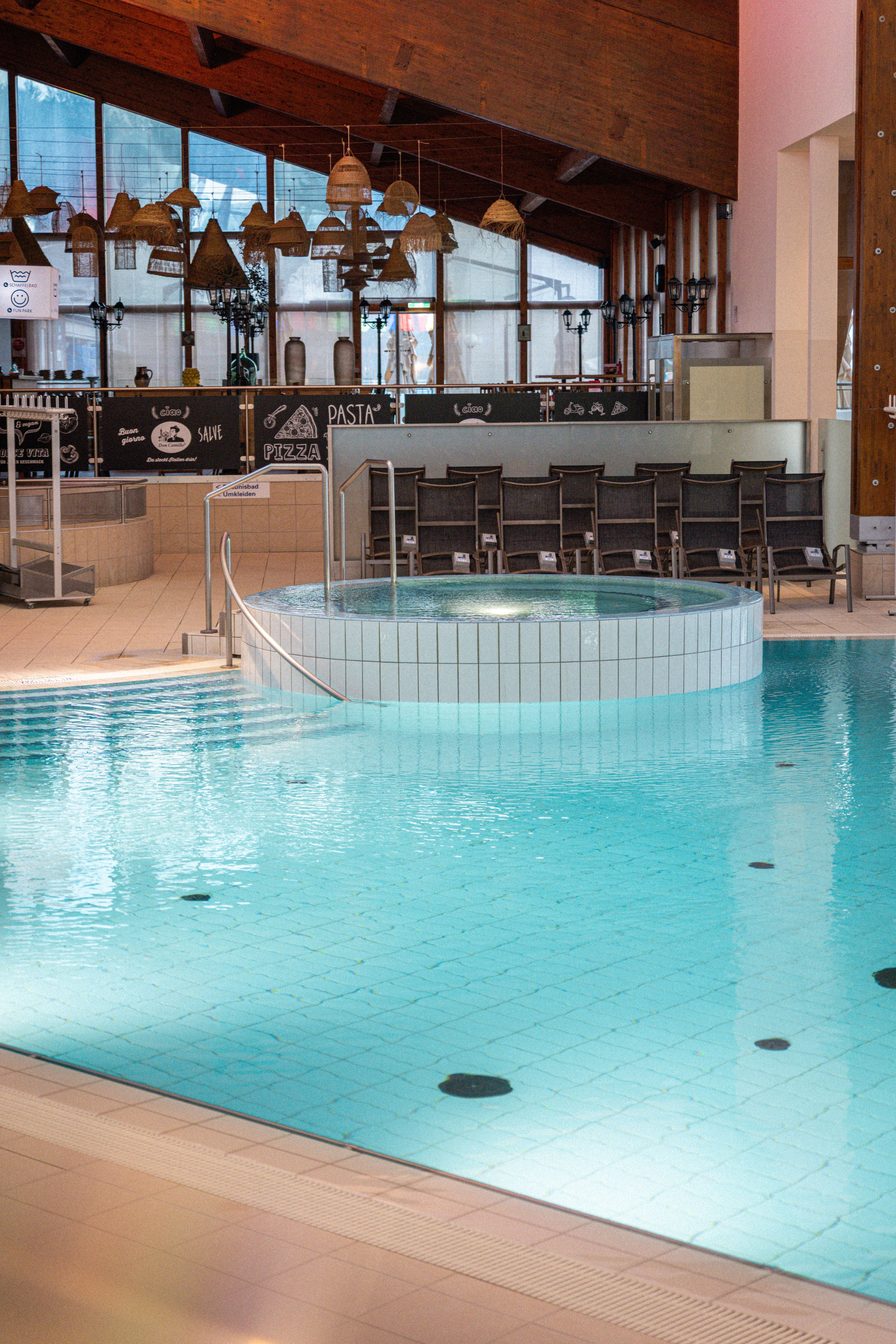 Innenansicht eines Schwimmbads mit rundem Whirlpool, Holzdecke und Bar im Hintergrund mit Stühlen und hängenden Lampen