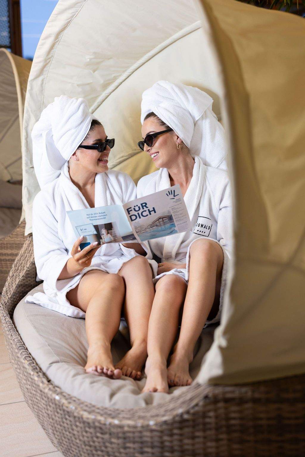 Two women sit in a cozy lounger and look together at the guest magazine “Für Dich” of the Loipersdorf thermal spa resort.
