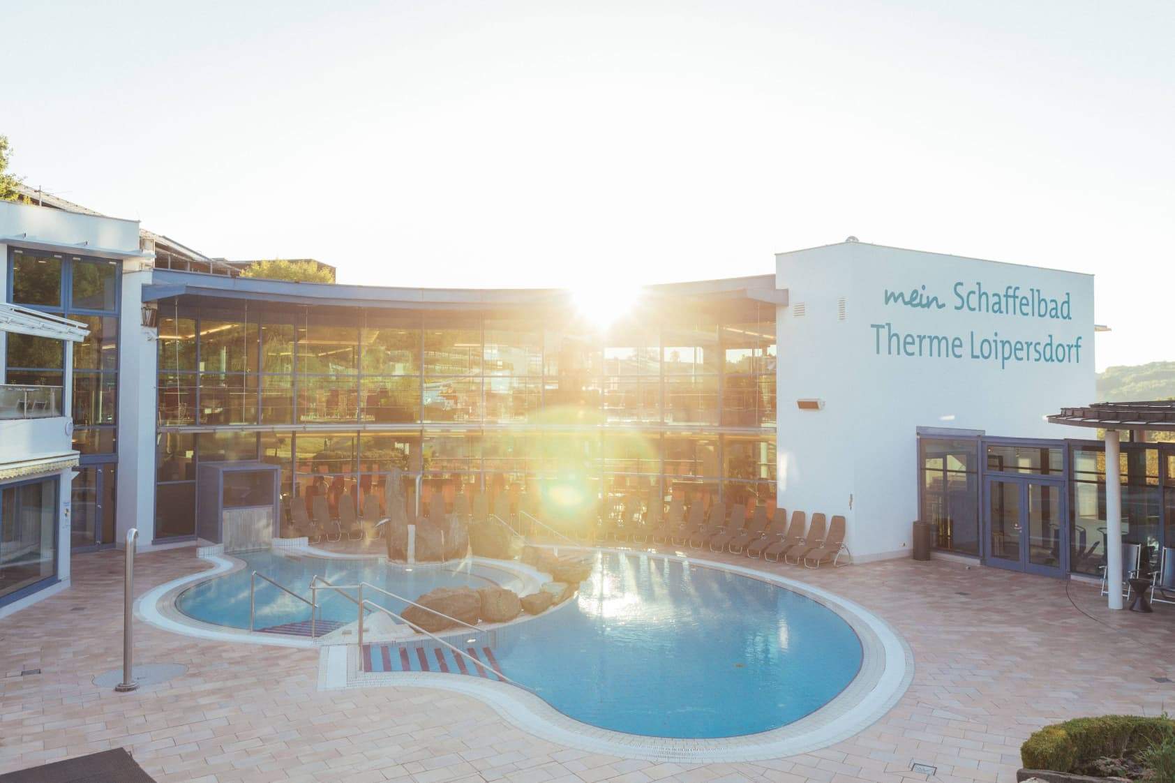 View of the thermal pool in the Schaffelbad. In the foreground the pool with the blue thermal water and in the background the architecture of the Schaffelbad with rays of sunshine.